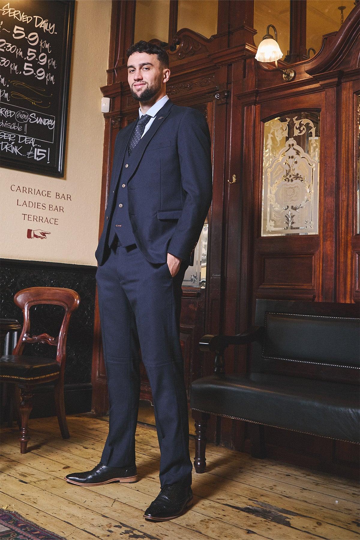 A man in the marcdarcy WHU Collection MAX Navy Three Piece Suit, as worn by Max Kilman, stands confidently in a vintage-style room with wood paneling, glass doors, antique chairs, and a chalkboard menu.