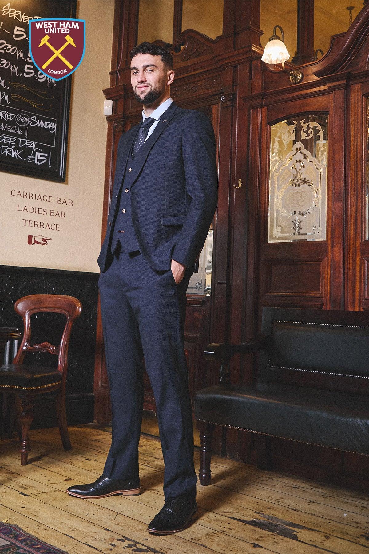A man in the marcdarcy WHU Collection - MAX Navy Three Piece Suit, as worn by Max Kilman, stands confidently in a vintage room with wooden panels and antique furnishings. The West Ham United logo appears in the top left corner.
