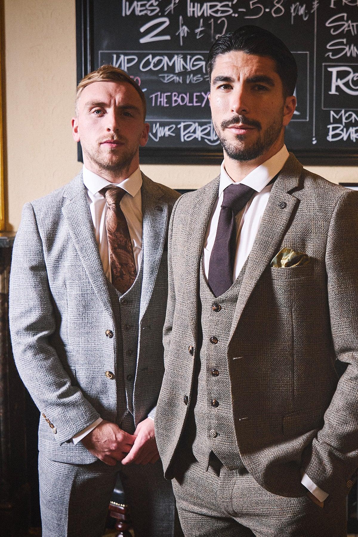 Two men in slim fit tweed suits stand before a chalkboard. On the left, one wears the marcdarcy WHU Collection - KRIS Silver Grey Tweed Suit as worn by Jarrod Bowen with a brown tie; on the right, a brown tweed suit with dark purple tie and green pocket square.