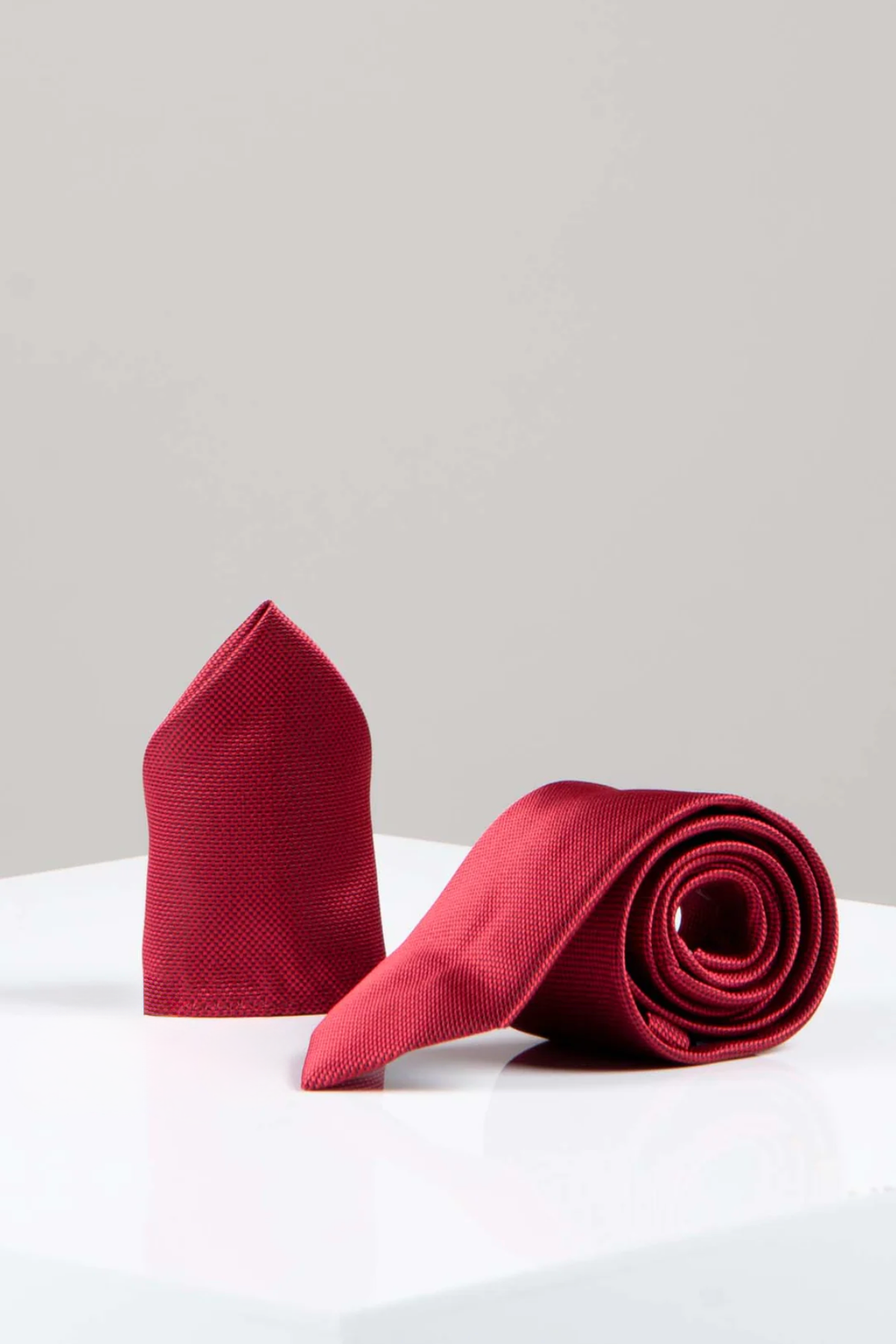 The Accessory TOBY Birdseye Tie and Pocket Square Set in Wine features an elegant rolled-up necktie with a subtle birdseye pattern and a matching pocket square, displayed on a white surface against a light gray background.