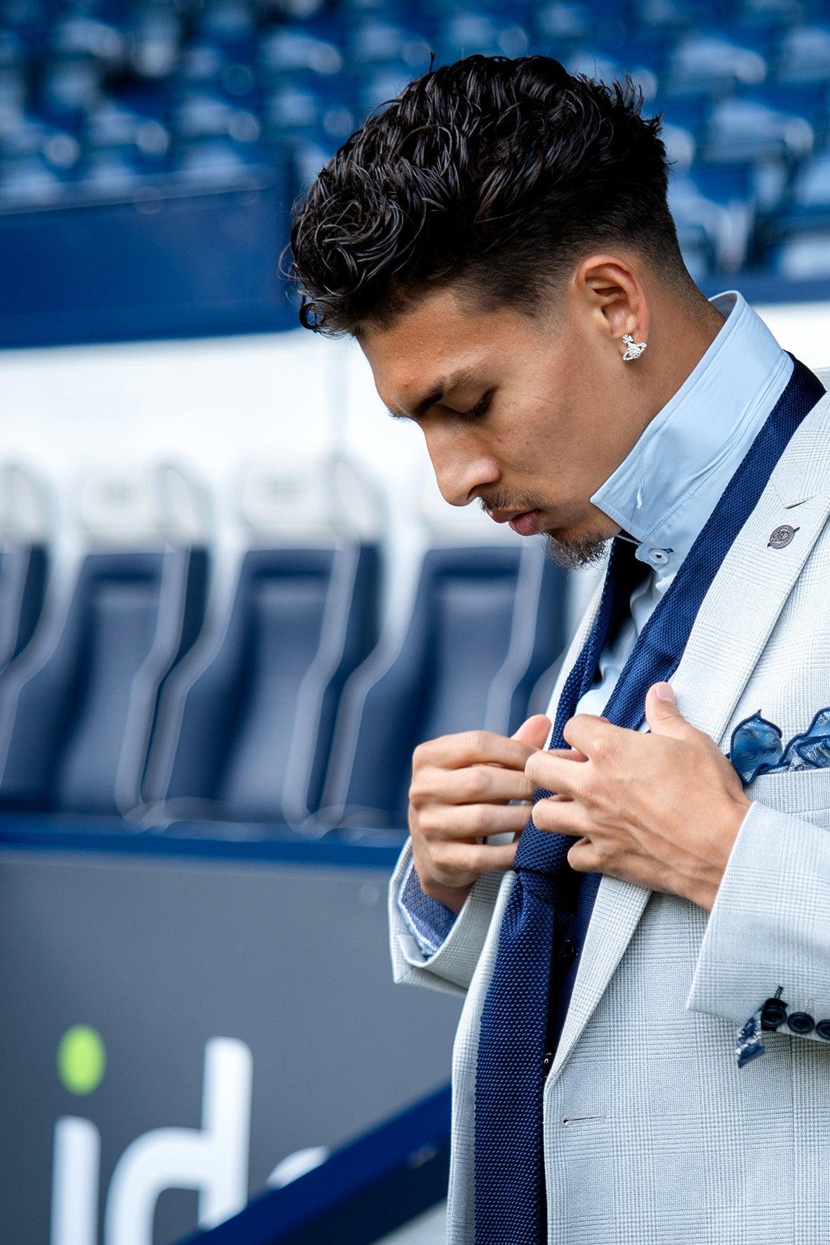 A man with dark, styled hair adjusts his tie while wearing the Marc Darcy WBA Collection - BROMLEY Stone Suit with a Kelvin Navy Waistcoat. Outdoors in front of empty stadium seats, he looks down with a focused expression.