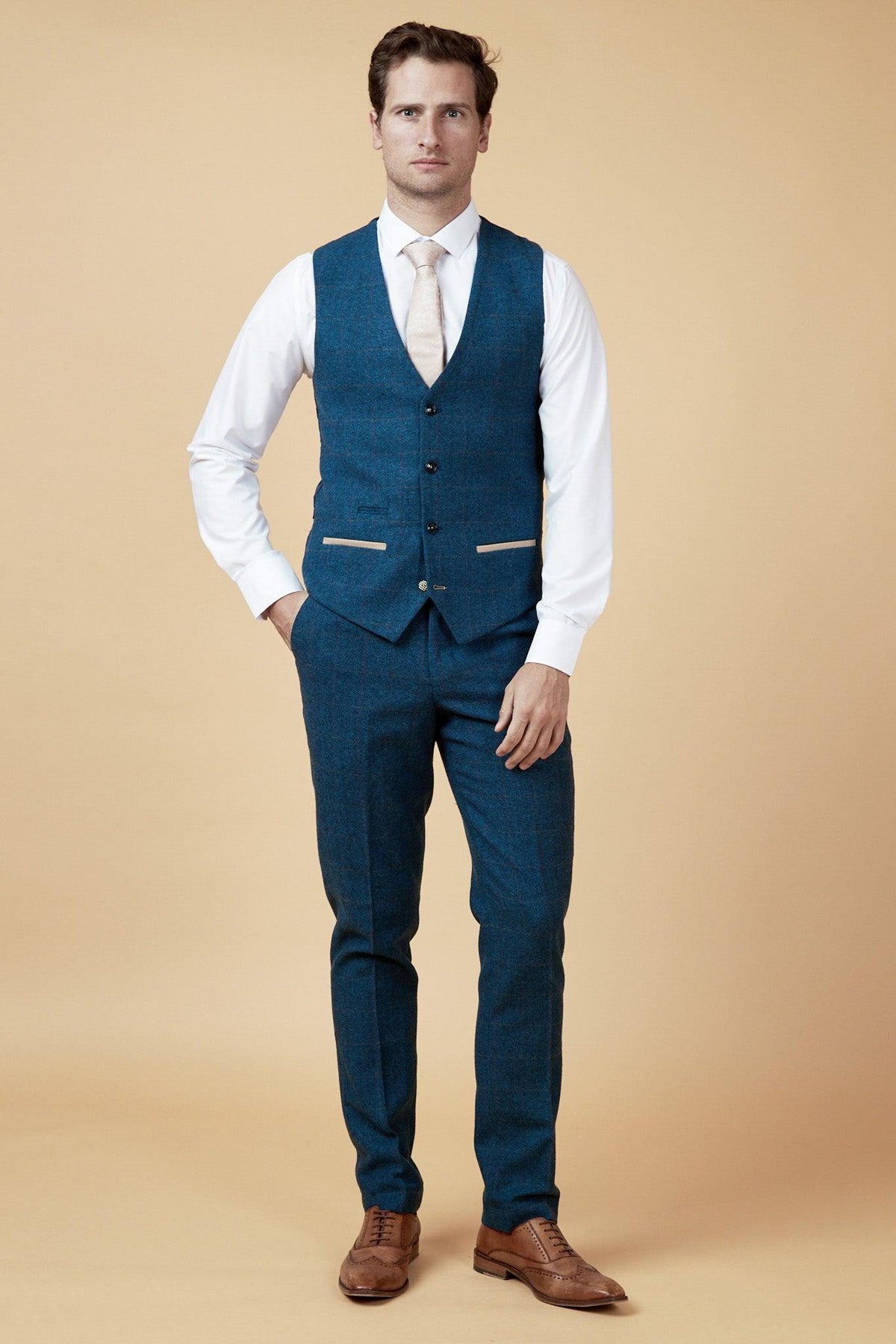A man wears the marcdarcy WHU Collection - TED Tan Tweed Suit with Dion Waistcoat, as worn by Lukasz Fabianski, standing against a tan background with his hand in his pocket.
