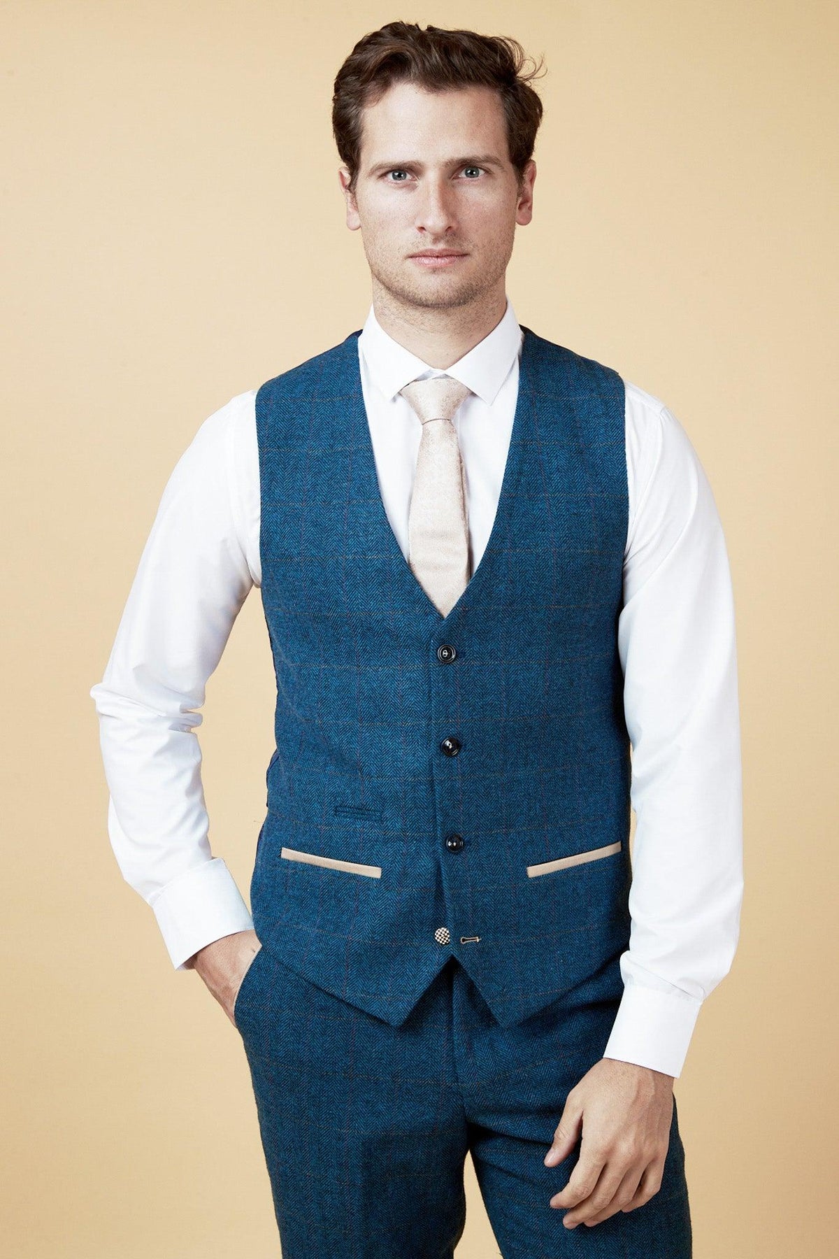 A man wears the marcdarcy WHU Collection TED Tan Tweed Suit with Dion Waistcoat, as worn by Lukasz Fabianski, paired with a white shirt and light tie, posing against a beige background with one hand in his pocket and a neutral expression.