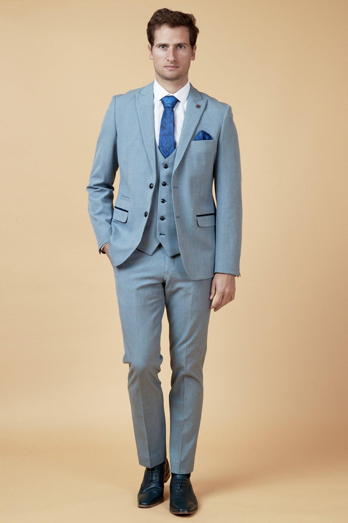 A man stands against a beige background, wearing the Marc Darcy WBA Collection - BROMLEY sky blue three-piece suit as worn by Brandon Thomas-Asante, with a Prince of Wales check, white shirt, blue tie, and matching pocket square.