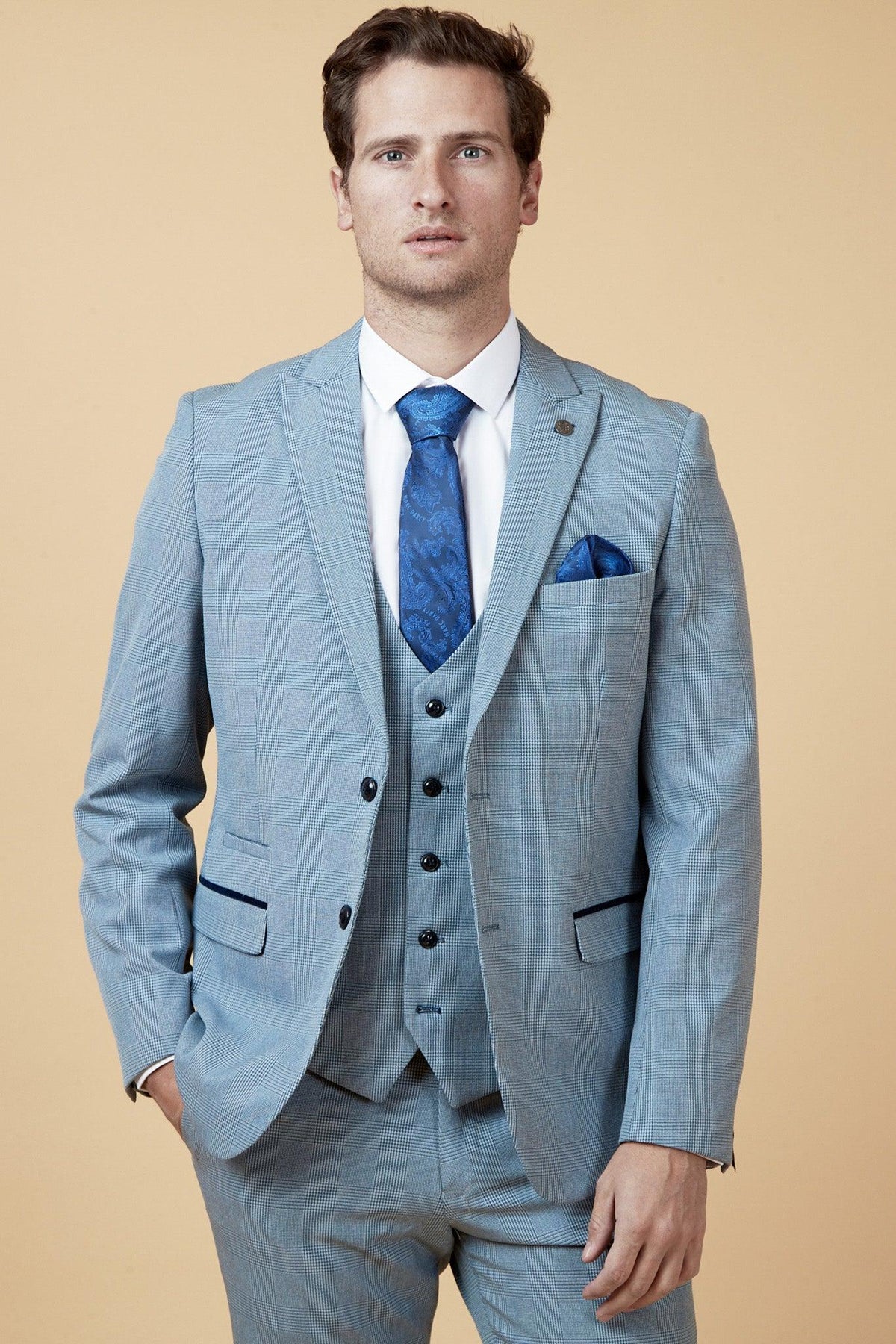 A man wears the Marc Darcy WBA Collection - BROMLEY Suit as worn by Brandon Thomas-Asante, featuring a sky blue three-piece design with matching tie and pocket square, standing against a beige background with one hand in his pocket.
