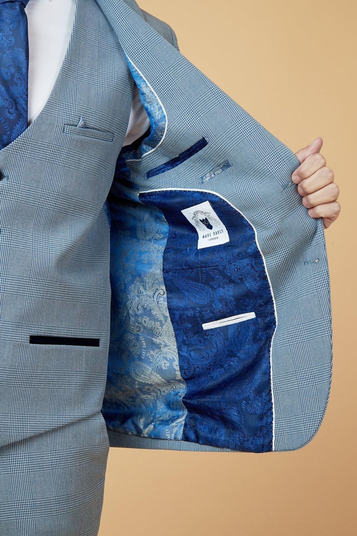 Calum Lill as Joel Deering from Coronation Street wears a Marc Darcy BROMLEY Sky Blue Blazer with wine chinos, opening the jacket to show off its patterned lining and inside label over a white shirt on a beige background.