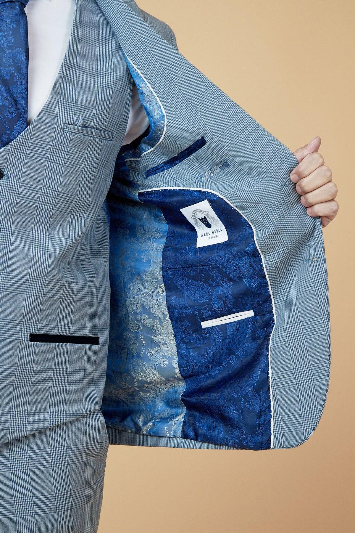 The WBA Collection - BROMLEY suit jacket by Marc Darcy, as worn by Brandon Thomas-Asante, opens to a vibrant blue paisley lining and interior pockets. Its exterior boasts a light grey Prince of Wales check, paired with a blue paisley tie.