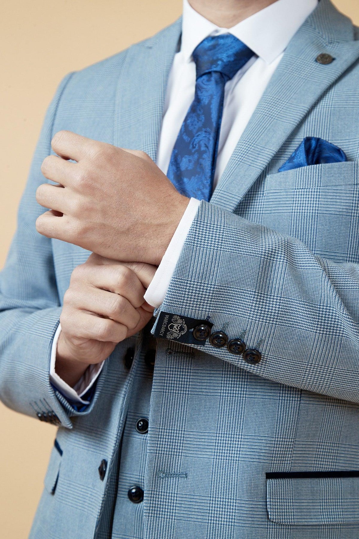 A man in Marc Darcy’s The WBA Collection - BROMLEY Suit as worn by Brandon Thomas-Asante, featuring a Prince of Wales check in sky blue, adjusts his cuff; he wears a white shirt, blue patterned tie, and shows a blue pocket square. Light beige background.