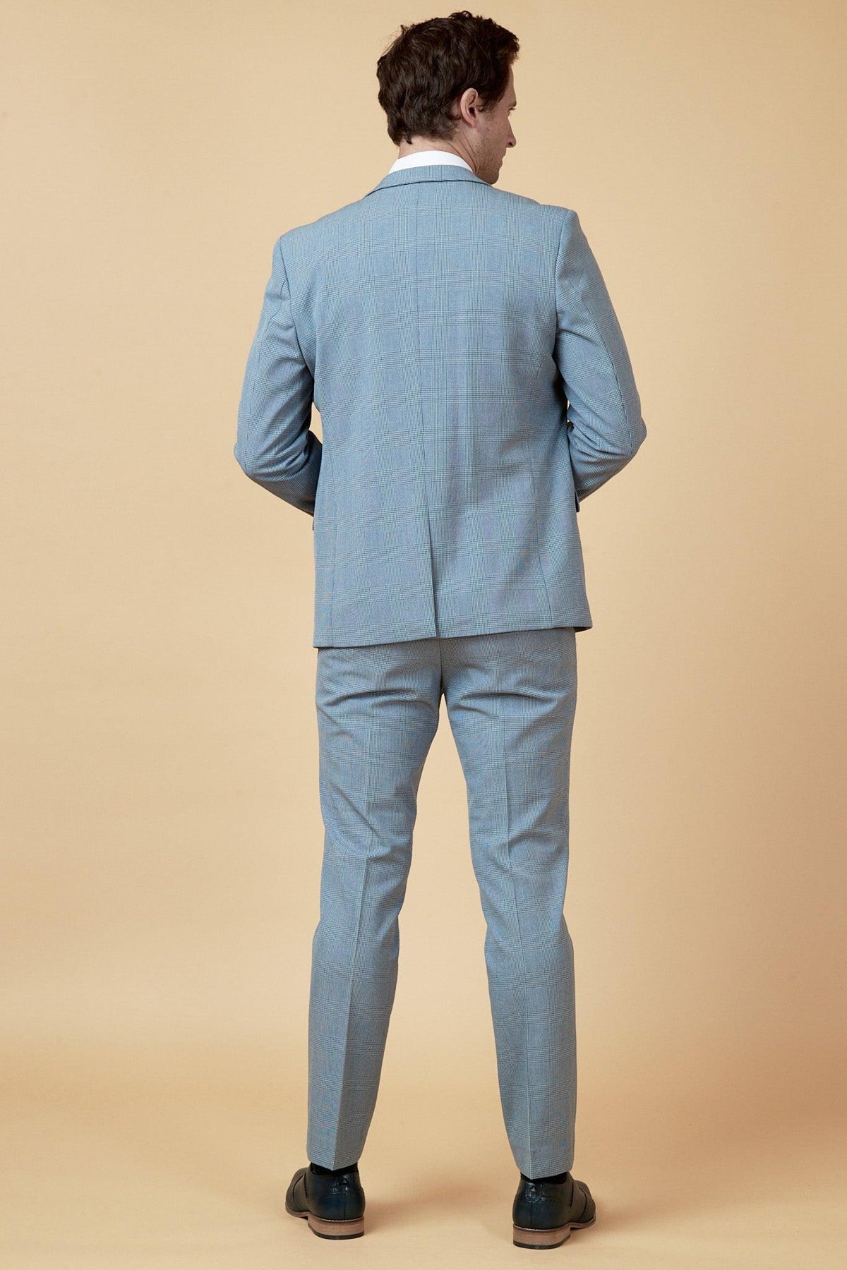 A man faces away from the camera, wearing the Marc Darcy WBA Collection - BROMLEY Suit as worn by Brandon Thomas-Asante, paired with black dress shoes against a plain beige background.