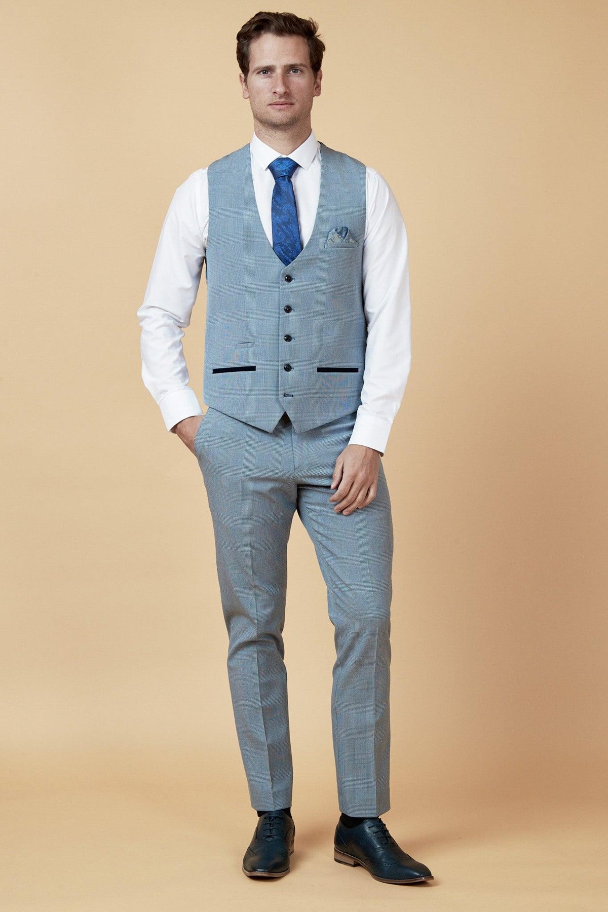 A man with short brown hair and a neutral expression stands against a beige background wearing the Marc Darcy WBA Collection - BROMLEY suit as worn by Brandon Thomas-Asante, styled with a white shirt, blue tie, pocket square, vest, and black shoes.