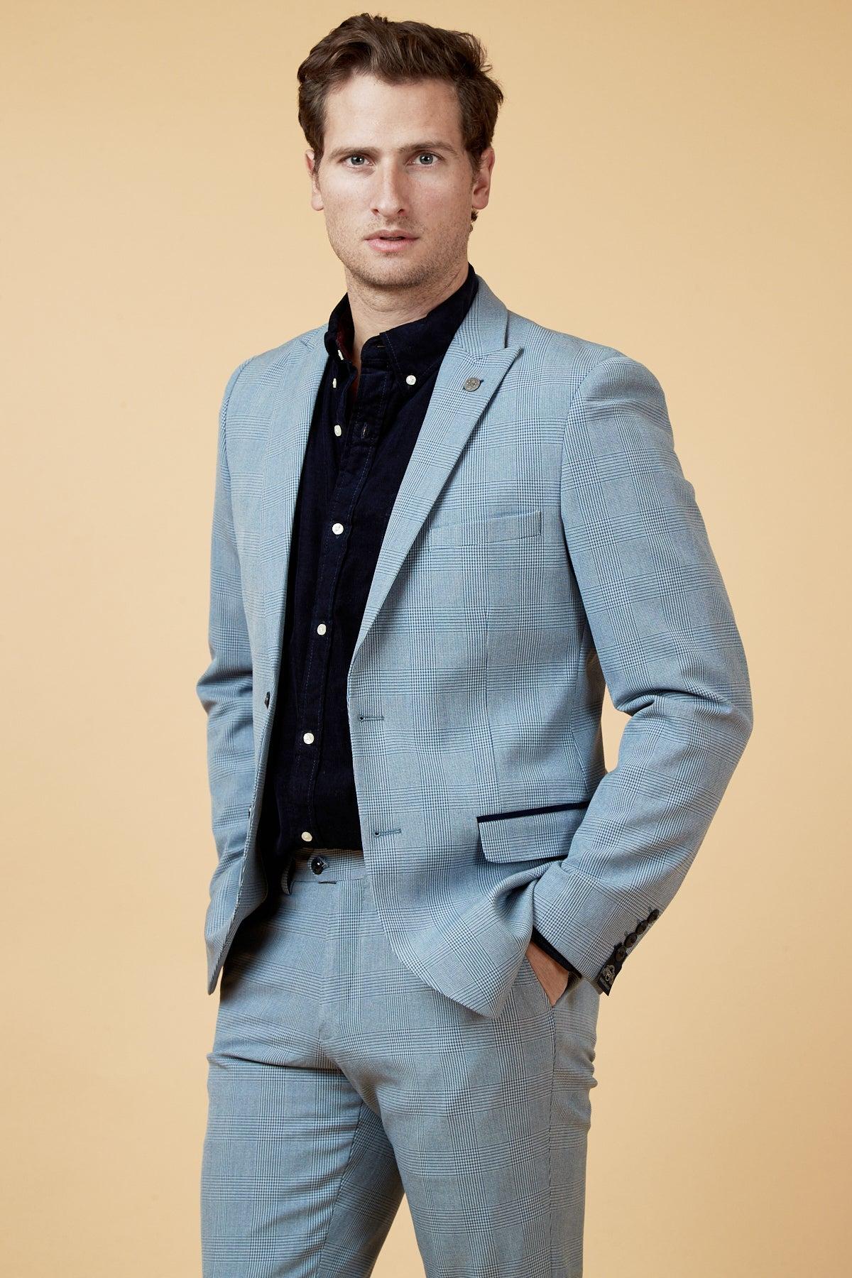 Coronation Streets Joel Deering (Calum Lill) models the Marc Darcy BROMLEY Sky Blue Blazer paired with wine chinos, standing confidently against a beige background with one hand in his pocket.