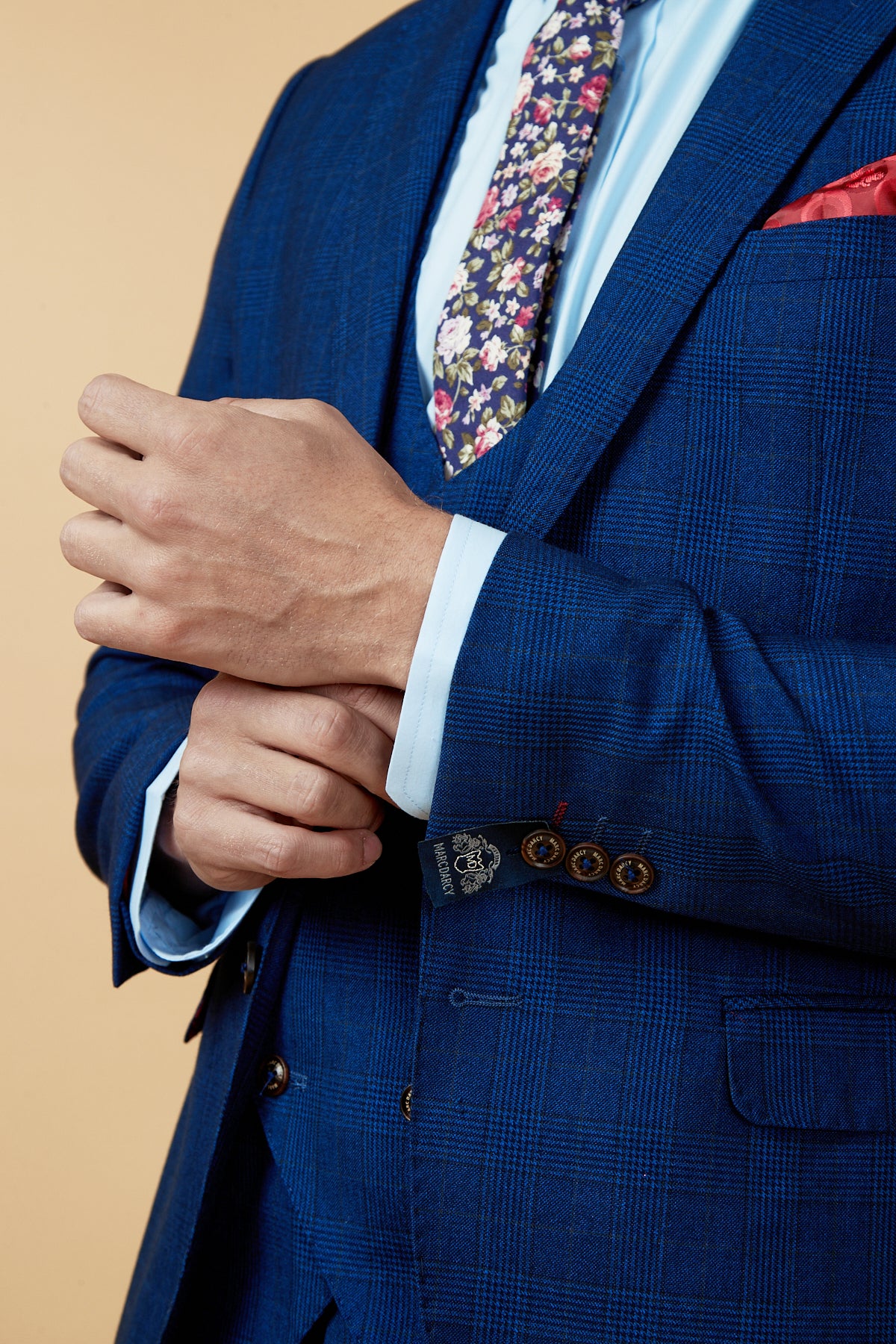 A man adjusts his marcdarcy GEORGE - Royal Blue Check Blazer, paired with a light blue shirt, floral tie, and red pocket square—a sharp choice for any work wardrobe.