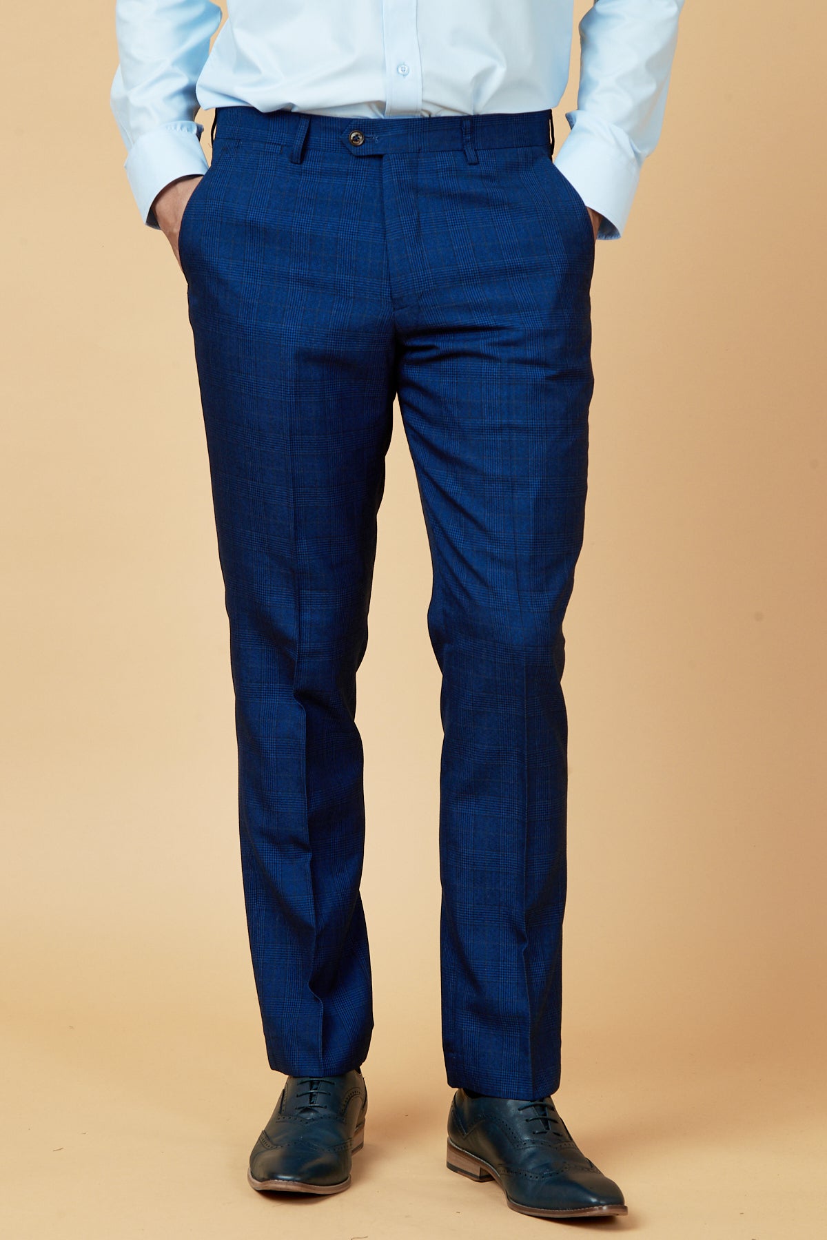 A person wearing marcdarcy GEORGE Royal Blue Check Trousers, a light blue dress shirt, and dark blue dress shoes stands with hands in pockets against a beige background.