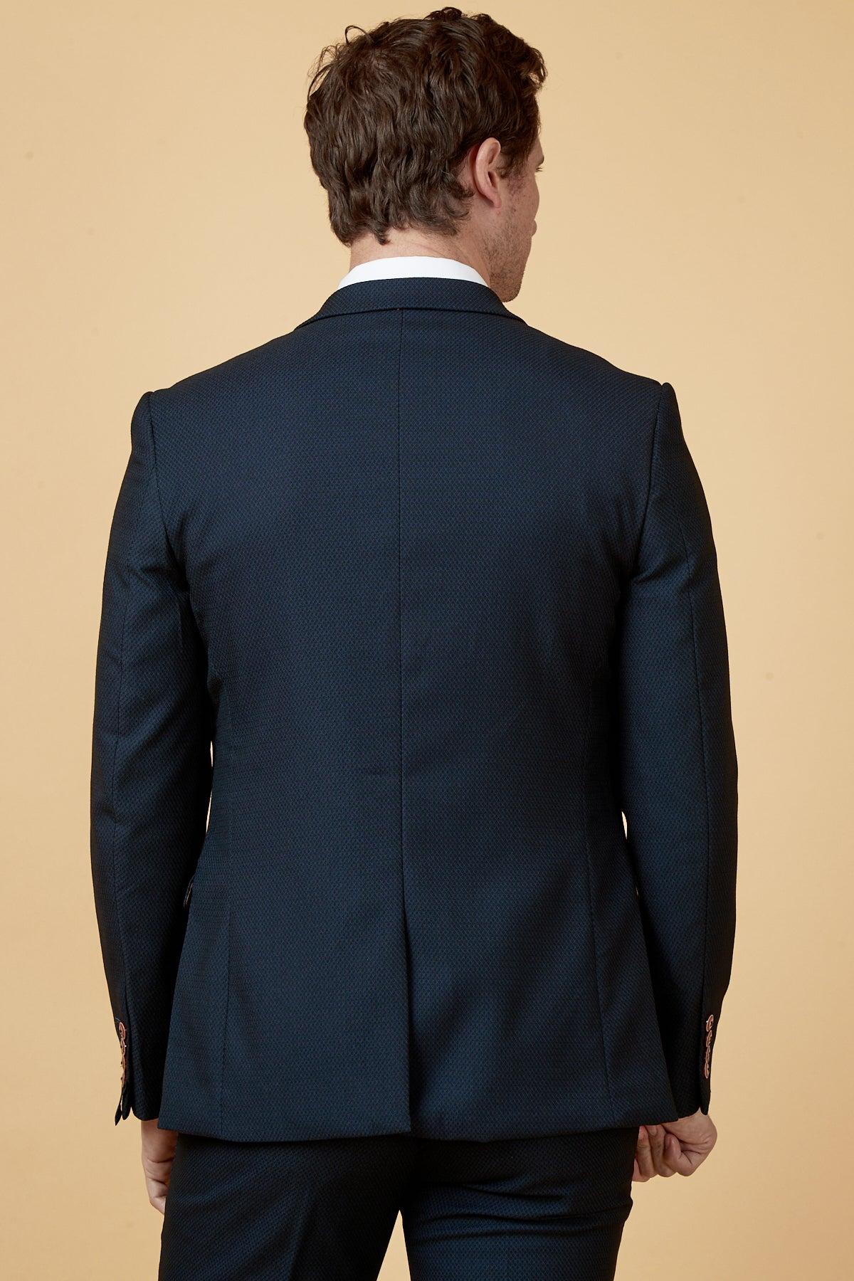 Dwight McNeil models The Everton Collection Max Navy Suit by marcdarcy, standing with his back to the camera against a plain beige background, showcasing the sharp peak lapel design.