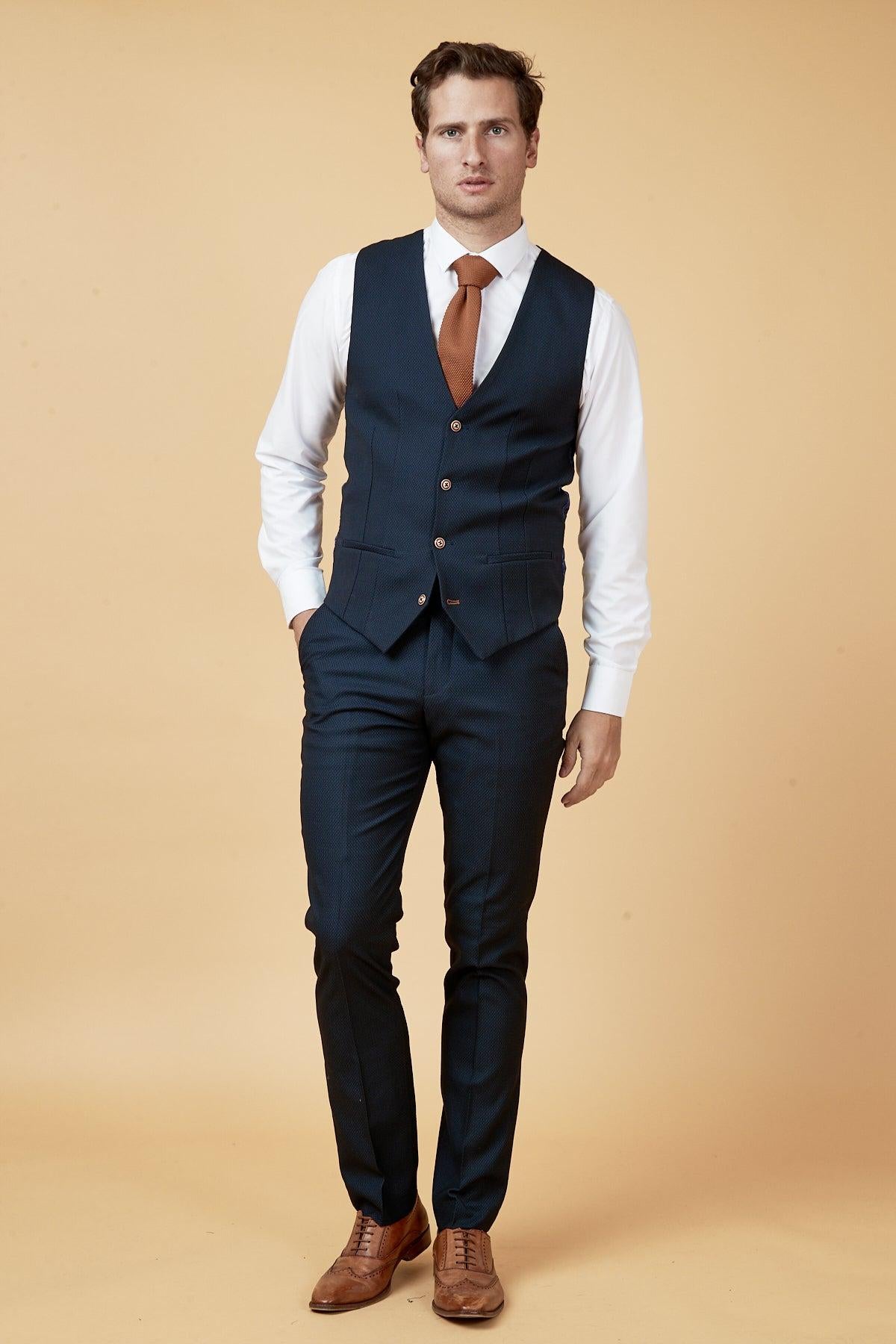 Dwight McNeil models The Everton Collection Max Navy Suit by marcdarcy, styled slim-fit with a white shirt, rust tie, and brown dress shoes. He stands against a beige backdrop with one hand in his pocket, looking at the camera.