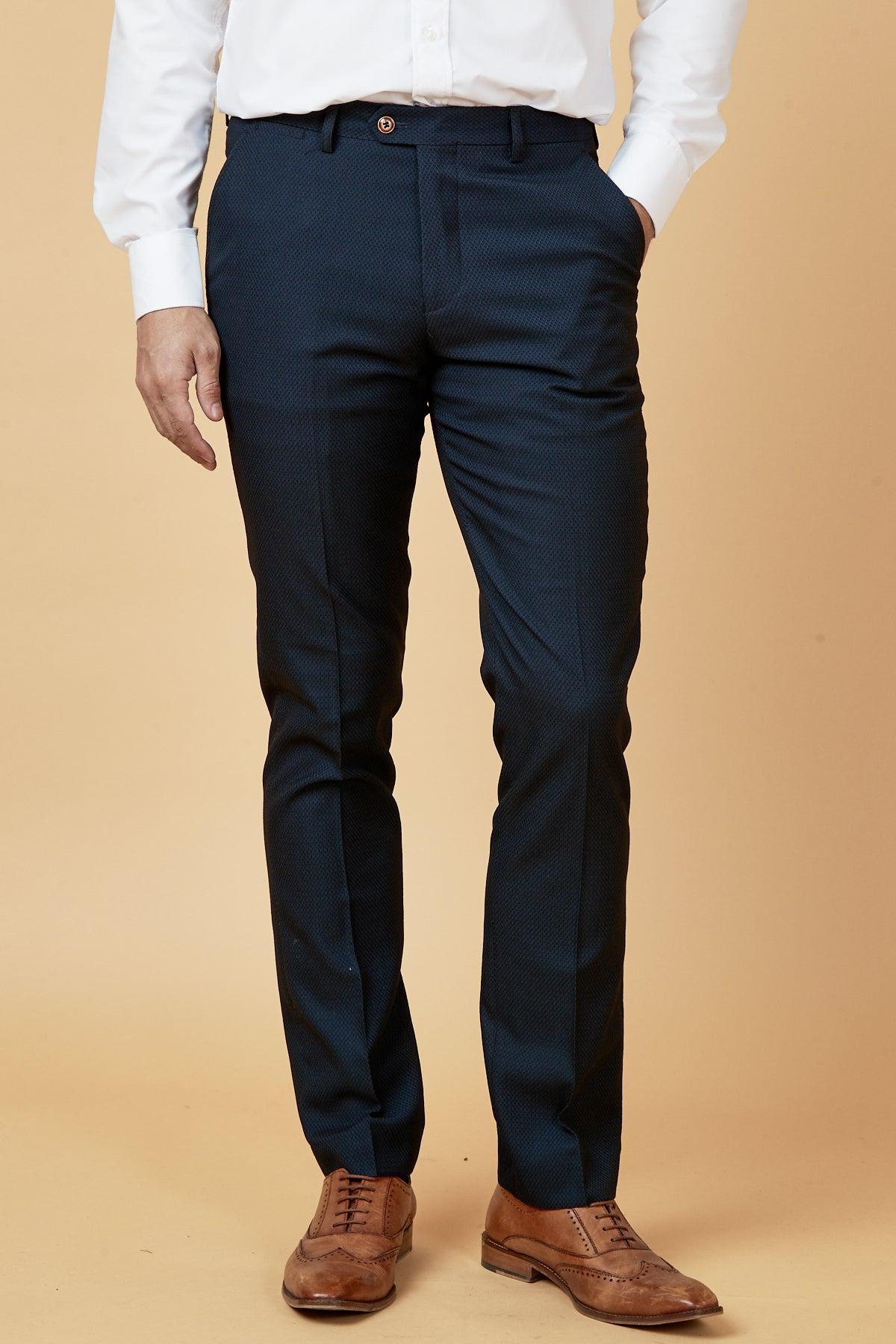 Dwight McNeil wears The Everton Collection Max Navy Suit by marcdarcy with a white shirt and brown leather shoes, standing against a beige background and showcasing a slim fit style with one hand in his pocket.