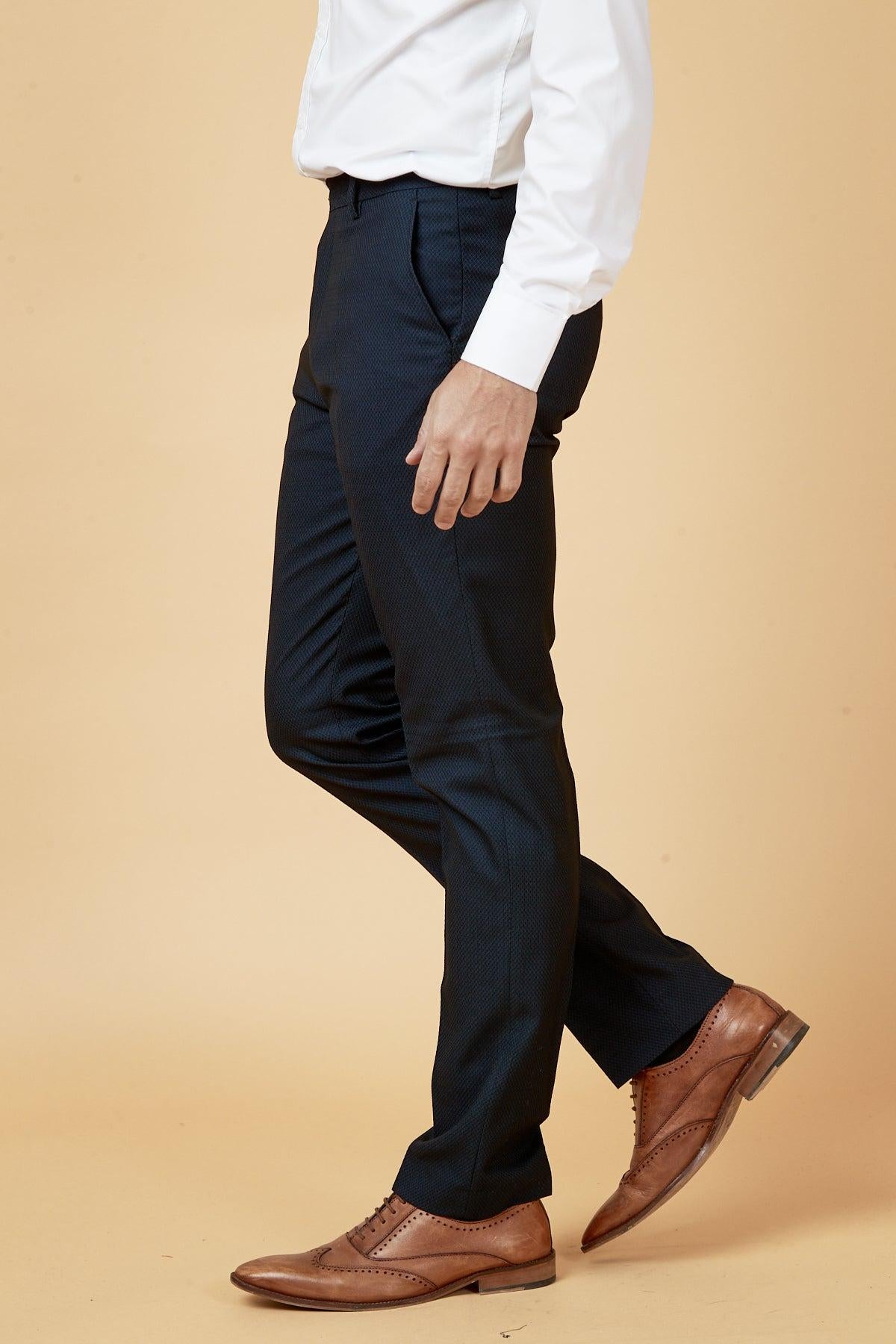A man models The Everton Collections Max Navy Suit by marcdarcy, wearing a slim fit jacket, white dress shirt, and brown leather shoes against a beige background. The image captures him from the shoulders down.