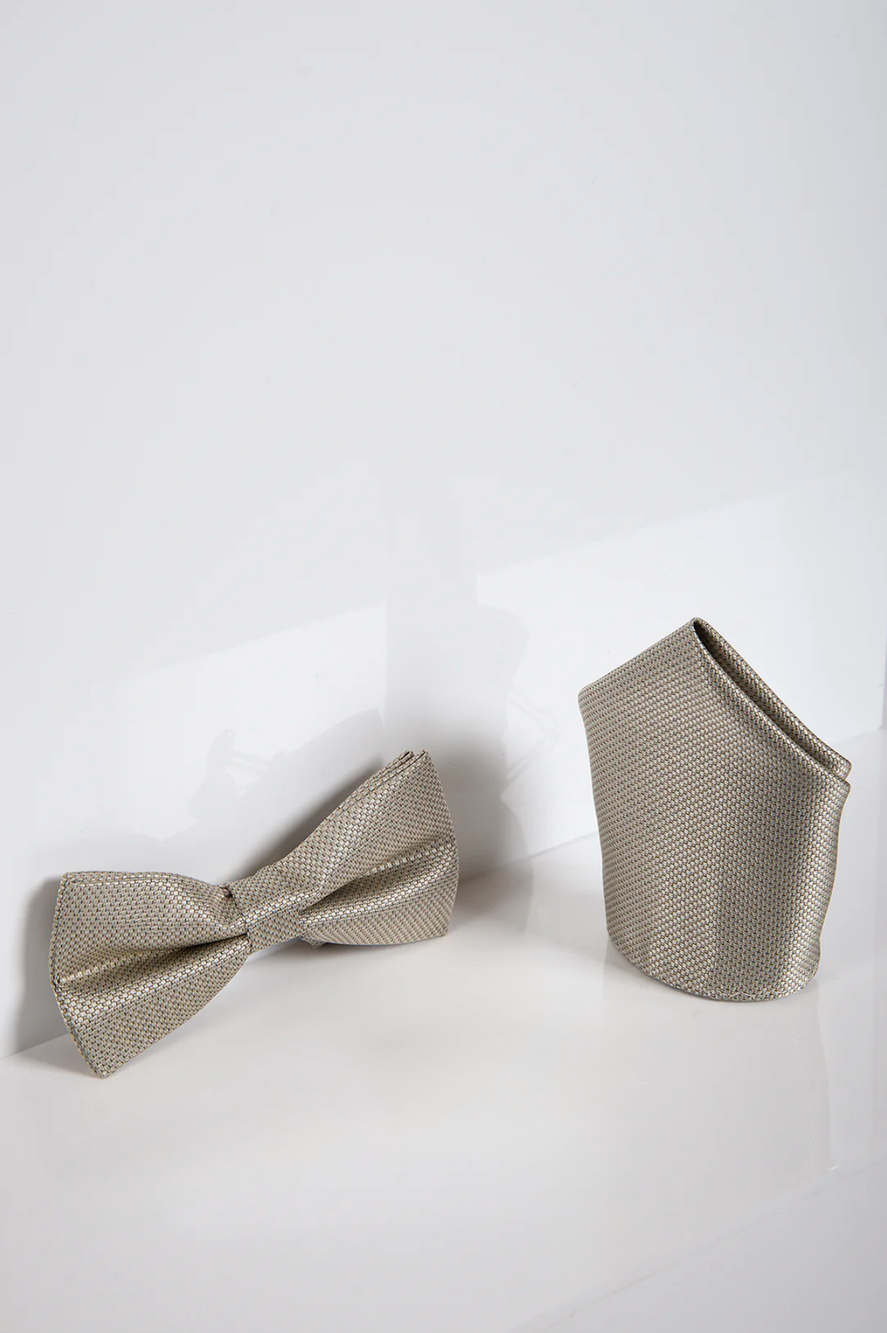 The Accessory TB17 Birdseye Print Bow Tie and Pocket Square Set in Tan is shown on a glossy white surface against a plain backdrop, offering a sophisticated and stylish accessory duo.