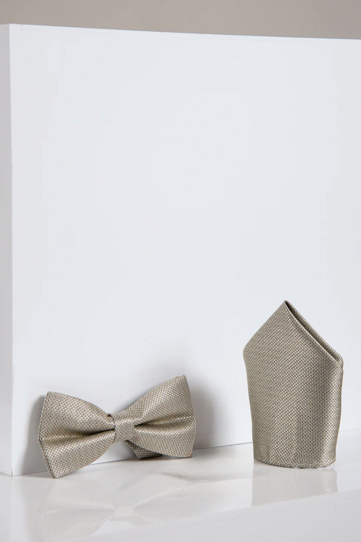 A TB17 Birdseye Print Bow Tie and Pocket Square Set in Tan by Accessory rests pre-tied and folded on a white reflective surface before a white block.