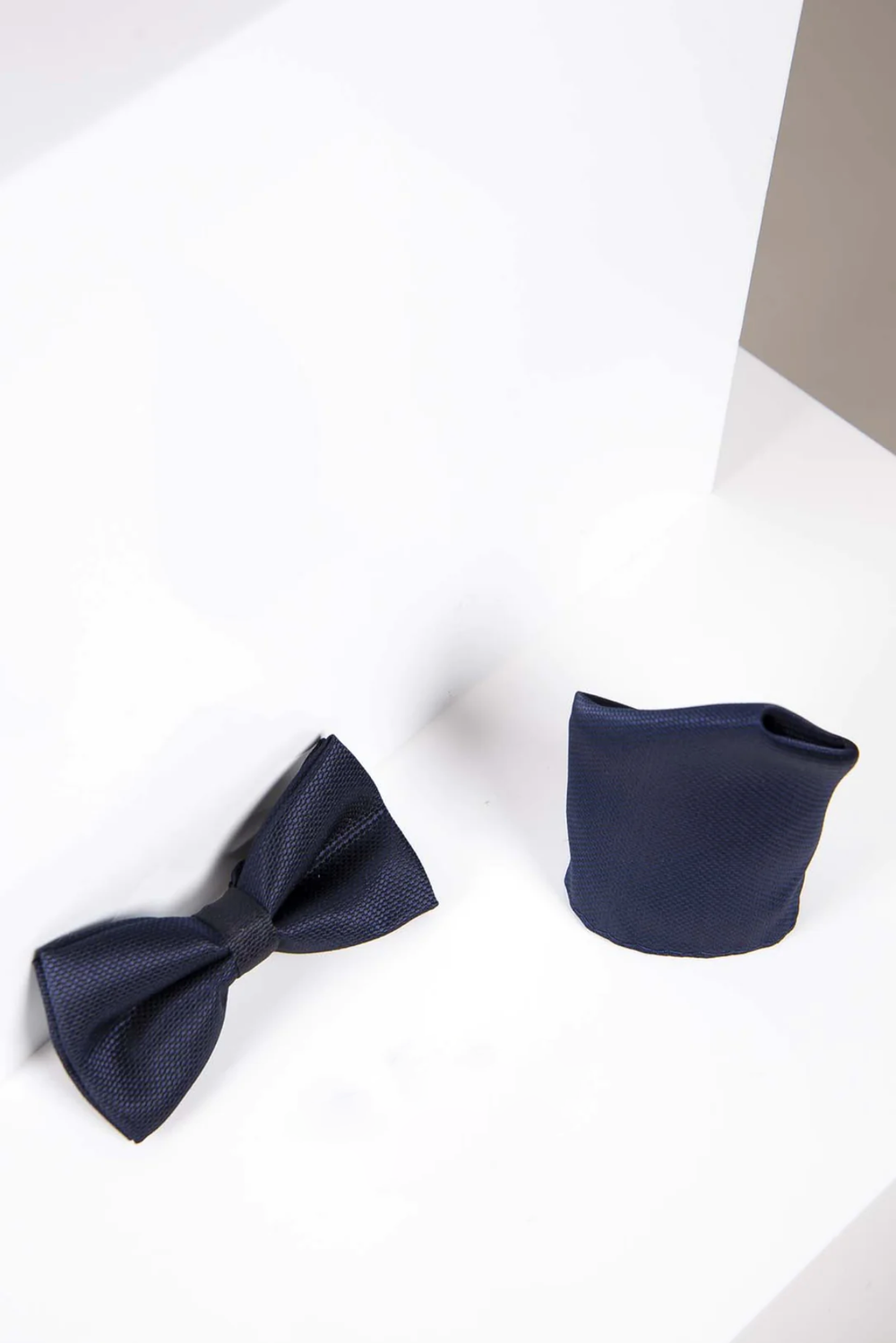 The Accessory TB17 Birdseye Print Bow Tie and Pocket Square Set in Navy features a textured bow tie with adjustable neck strap and matching pocket square, shown on a white, angled-edged surface.