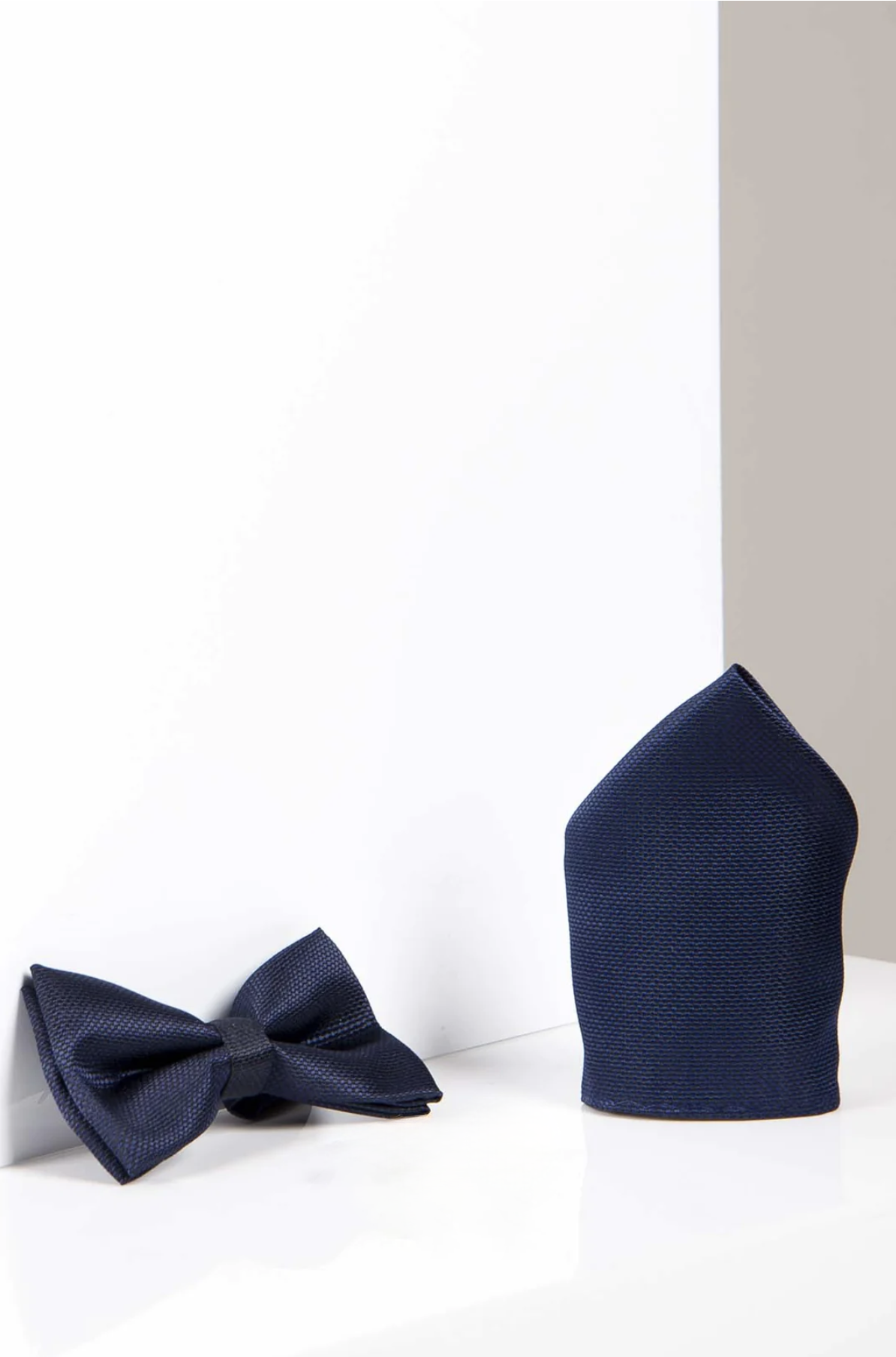 The Accessory TB17 Birdseye Print Bow Tie and Pocket Square Set in Navy features a textured bow tie with an adjustable strap and a matching folded pocket square, shown on a white and light gray background.
