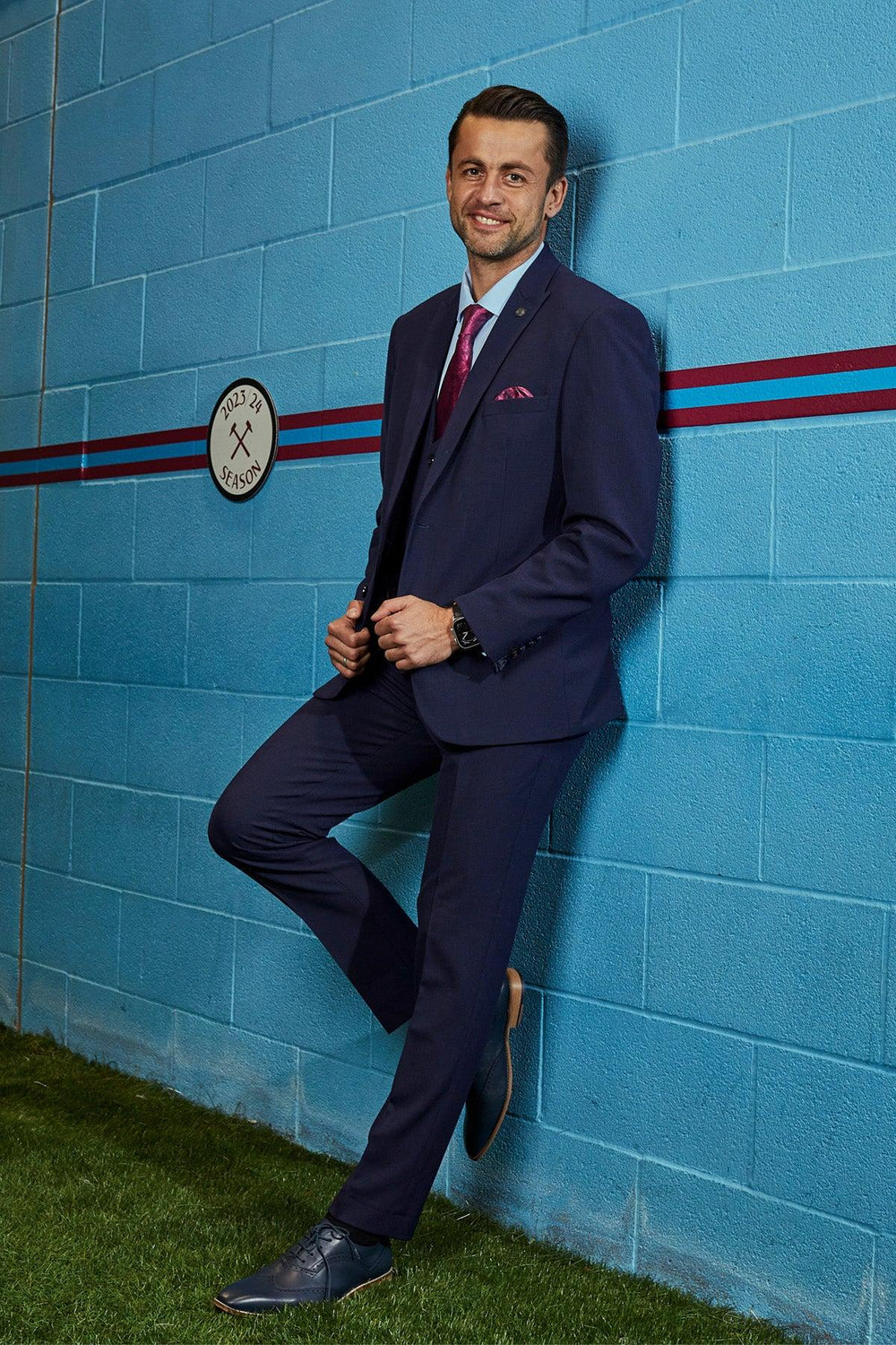 A man in The WHU Collection - BROMLEY Navy Check Three Piece Suit by Marc Darcy, as worn by Lukasz Fabianski, smiles at the camera, leaning against a blue wall with a West Ham United circular sign.
