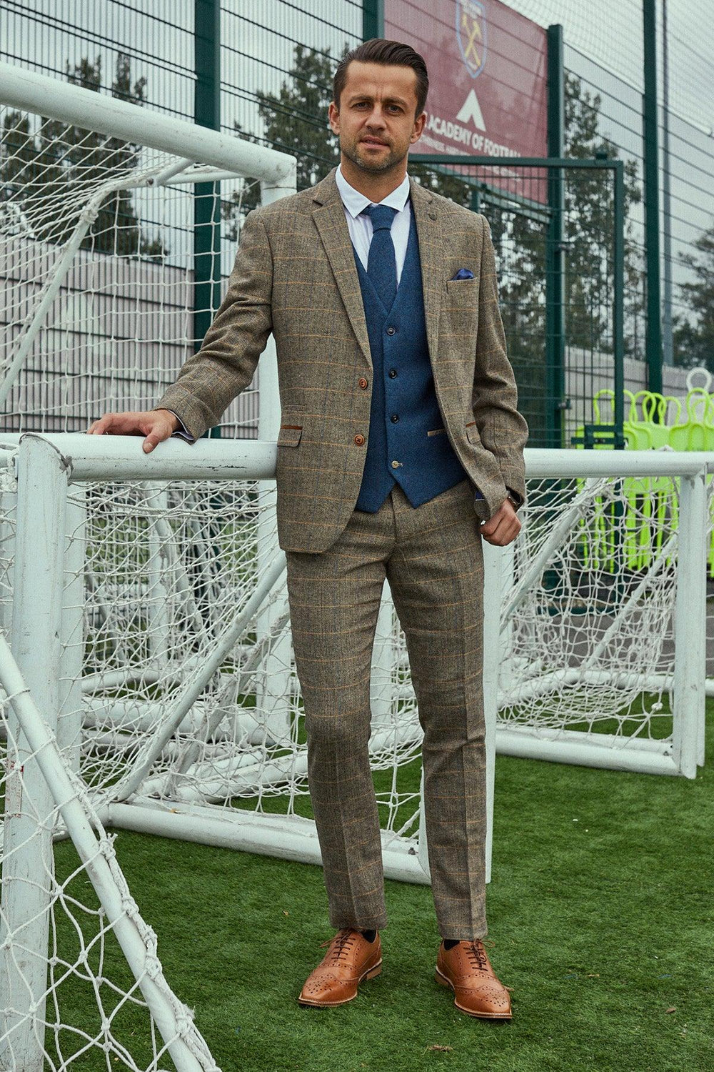 A man in the marcdarcy WHU Collection—TED Tan Tweed Suit with Dion Waistcoat as worn by Lukasz Fabianski—stands on artificial turf near a football goal, resting one hand on the net frame.