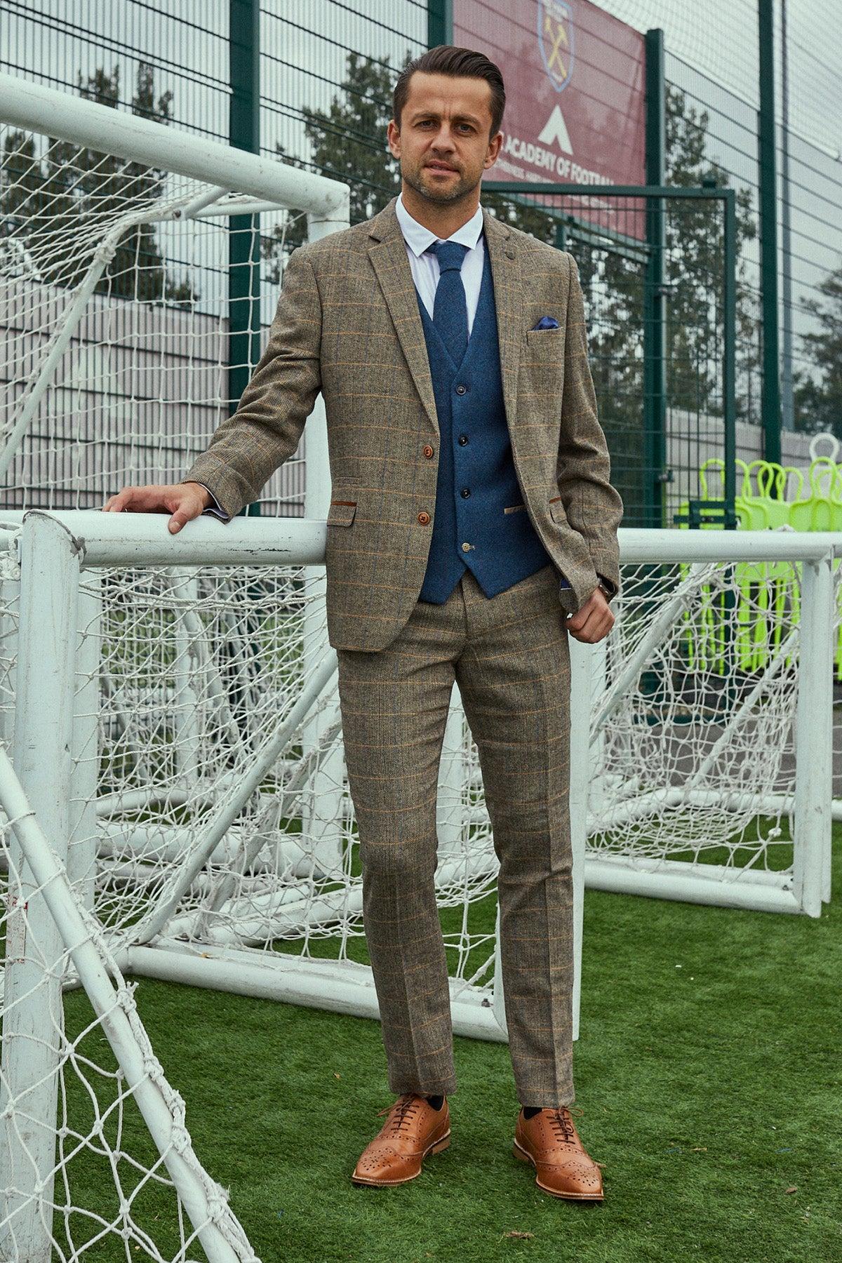 A man in the marcdarcy WHU Collection—TED Tan Tweed Suit with Dion Waistcoat as worn by Lukasz Fabianski—stands on artificial turf near a football goal, resting one hand on the net frame.