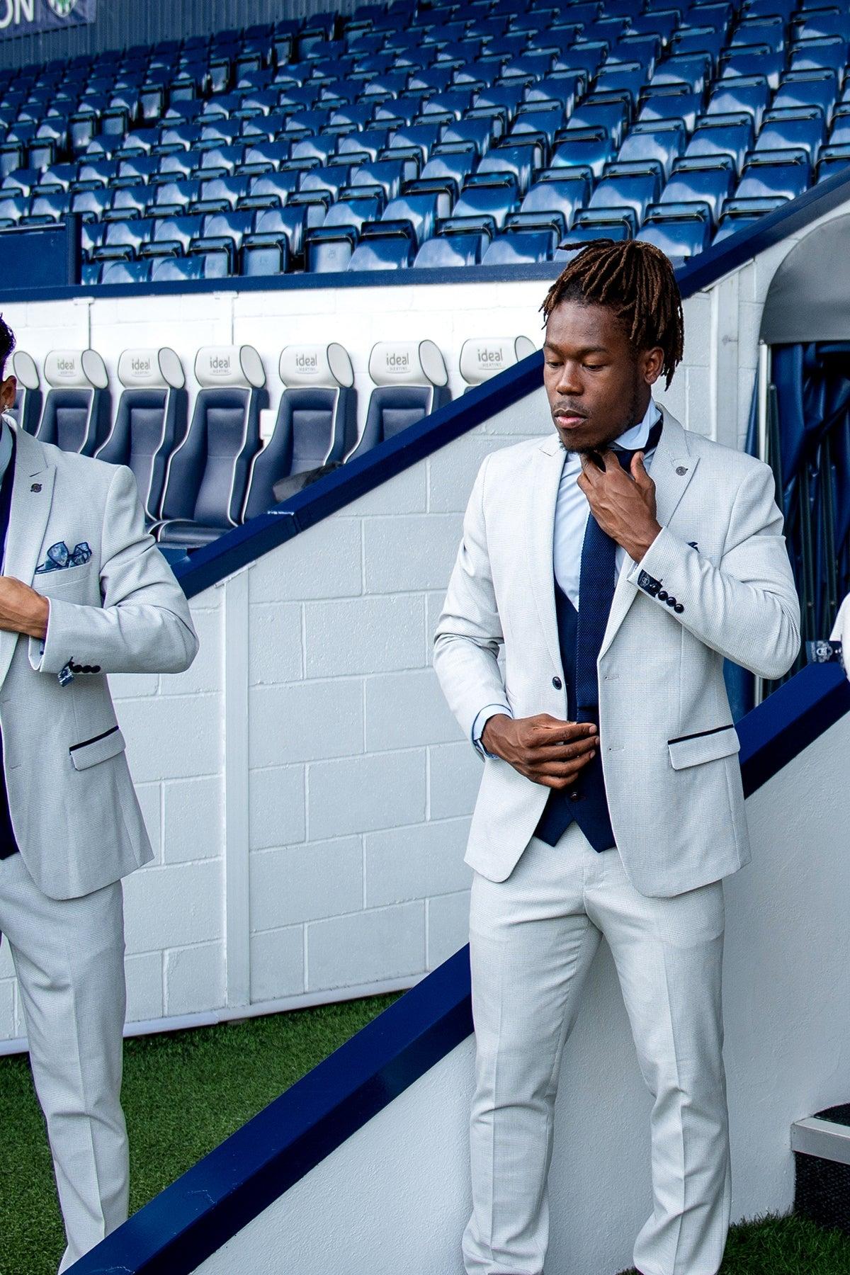 Two Black men wear Marc Darcy's WBA Collection—BROMLEY Stone Suits with Kelvin Navy Waistcoats—standing on a stadium walkway. The man on the right adjusts his tie.