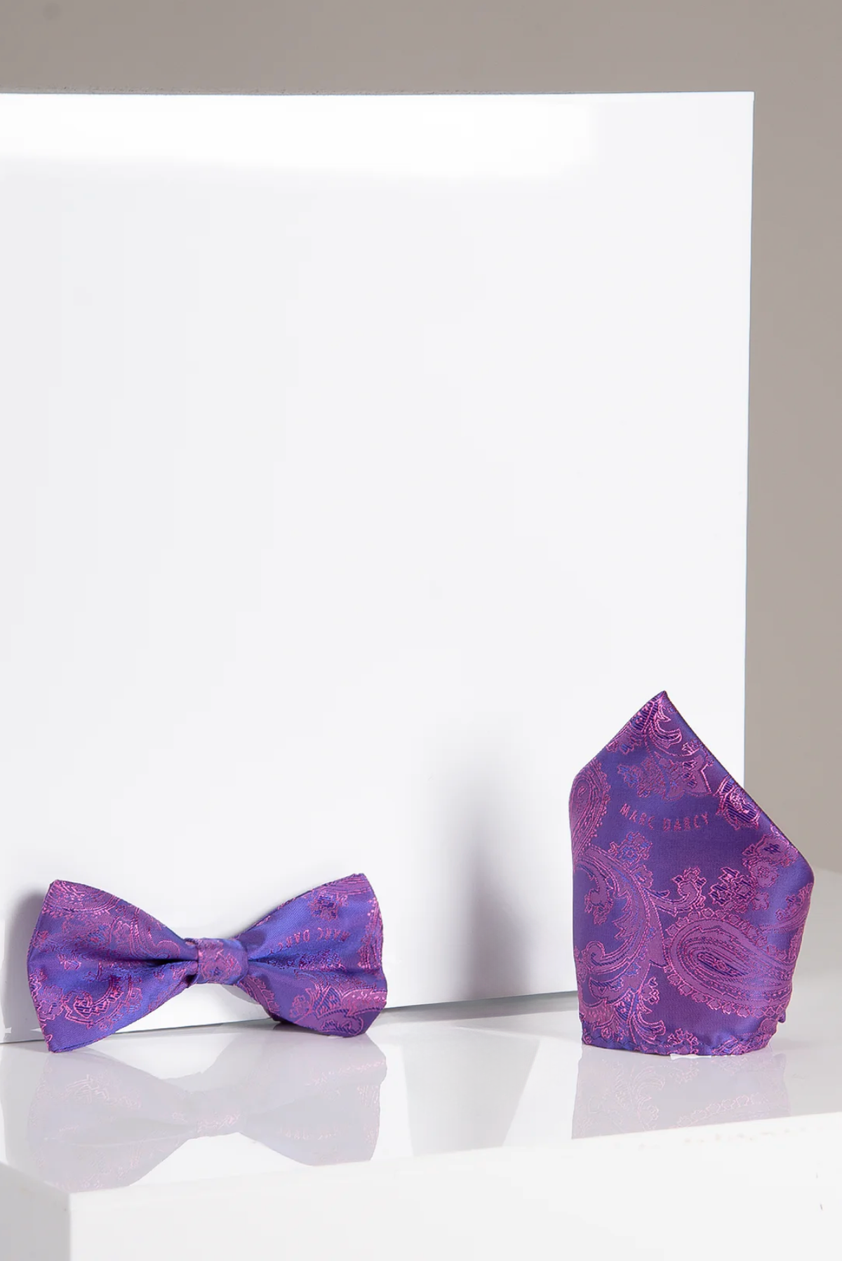 The Accessory MD PAISLEY Purple Paisley Bow Tie and Pocket Square Set is shown on a glossy white surface against a plain background.