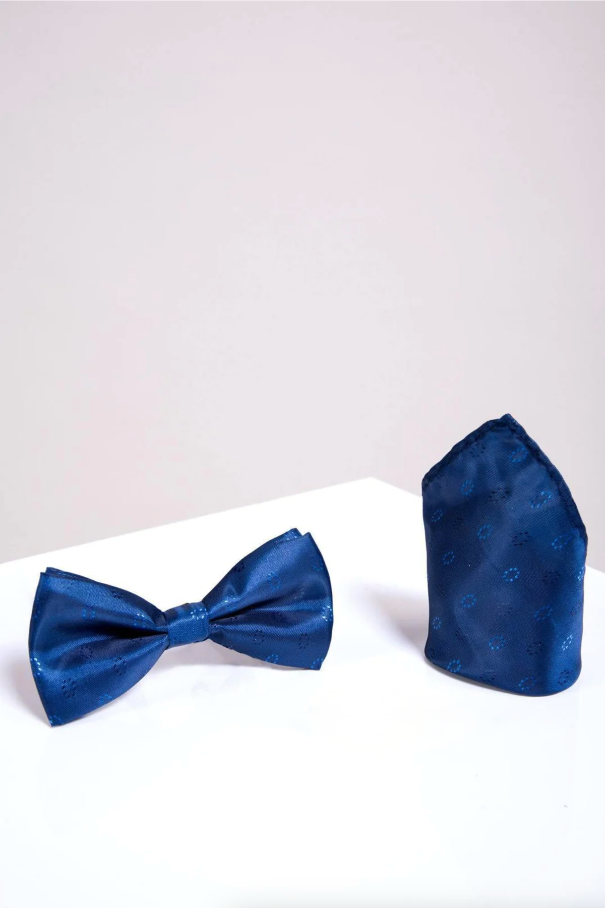 The BT CIRCLES - Blue Circle Print Bow Tie and Pocket Square Set by Accessory features a royal blue satin bow tie and matching pocket square with subtle circle prints, displayed on a white surface against a pale background.
