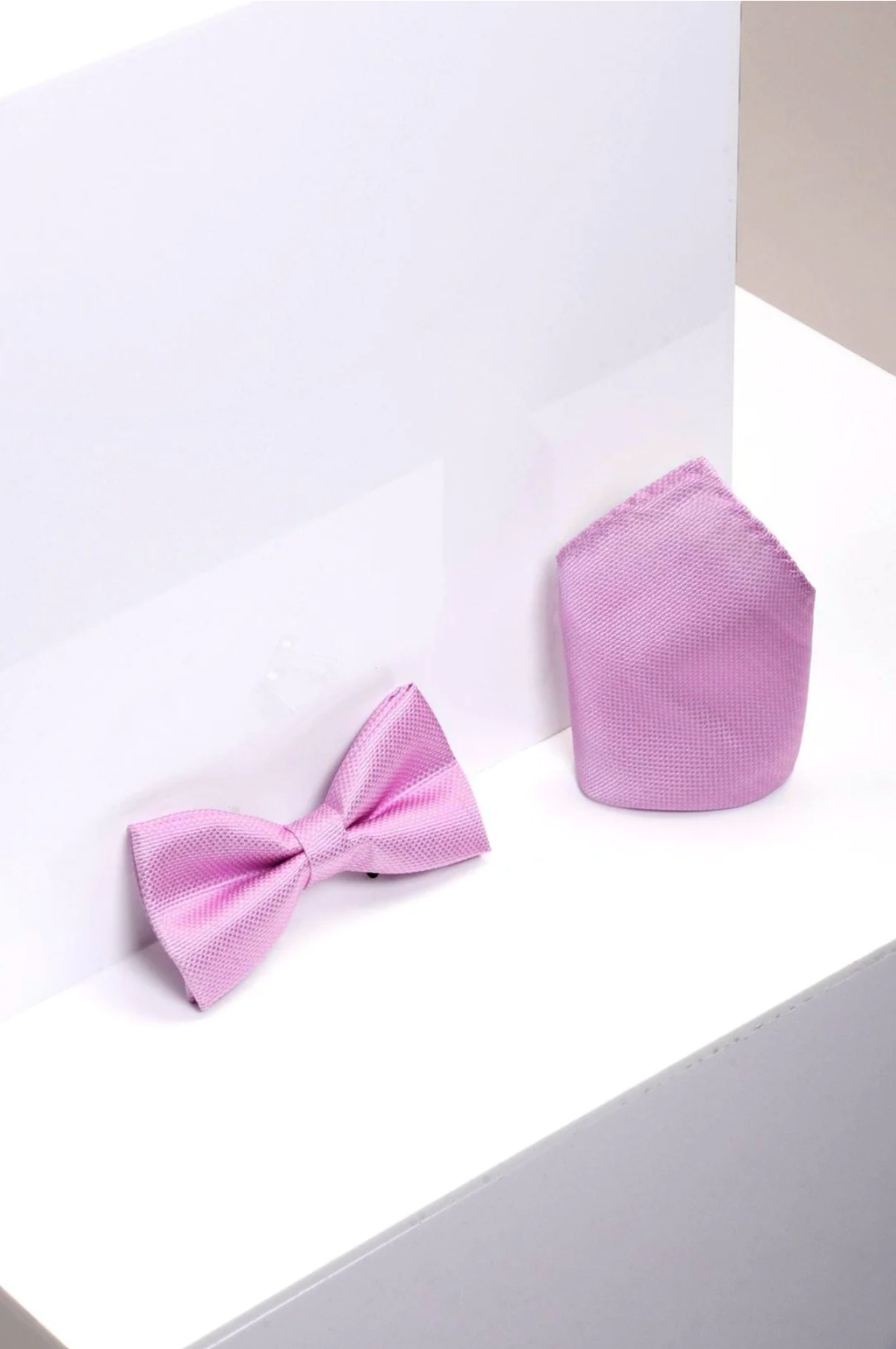 The Accessory TB17 Birdseye Print Bow Tie and Pocket Square Set in pink is showcased on a white geometric surface with a minimalist backdrop—an ideal, refined choice for wedding attire.