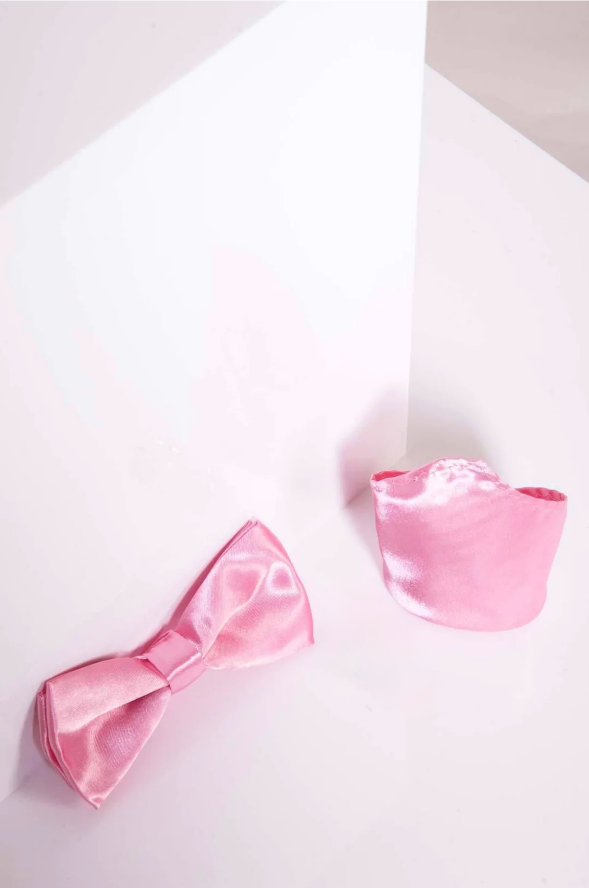 The Accessory SB - Satin Bow Tie and Pocket Square Set in Pink, shown on a glossy white surface with soft shadows, adds flair to formal wear with its classic satin style and matching pieces.