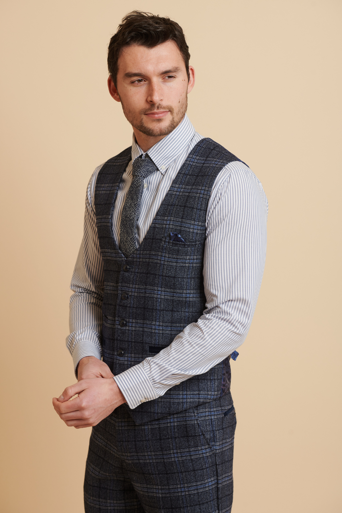 A man with short dark hair and a beard wears the Marc Darcy ANDY - Blue Single Breasted Check Waistcoat, styled with matching pants and a light striped shirt with tie, posing against a plain beige background.