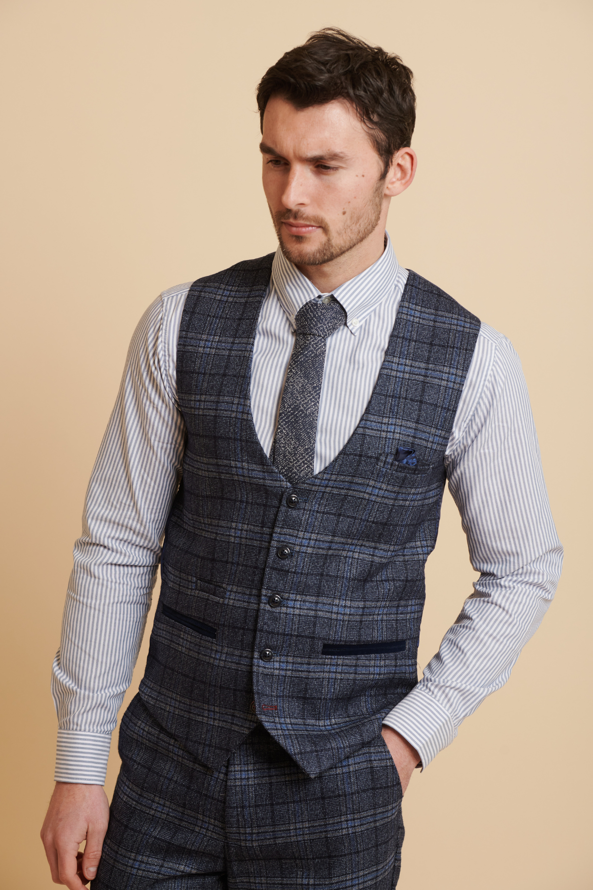 A man wears the Marc Darcy ANDY blue single breasted check waistcoat and trousers with a white and blue striped shirt and matching tie, standing against a beige background, looking slightly down with one hand in his pocket.
