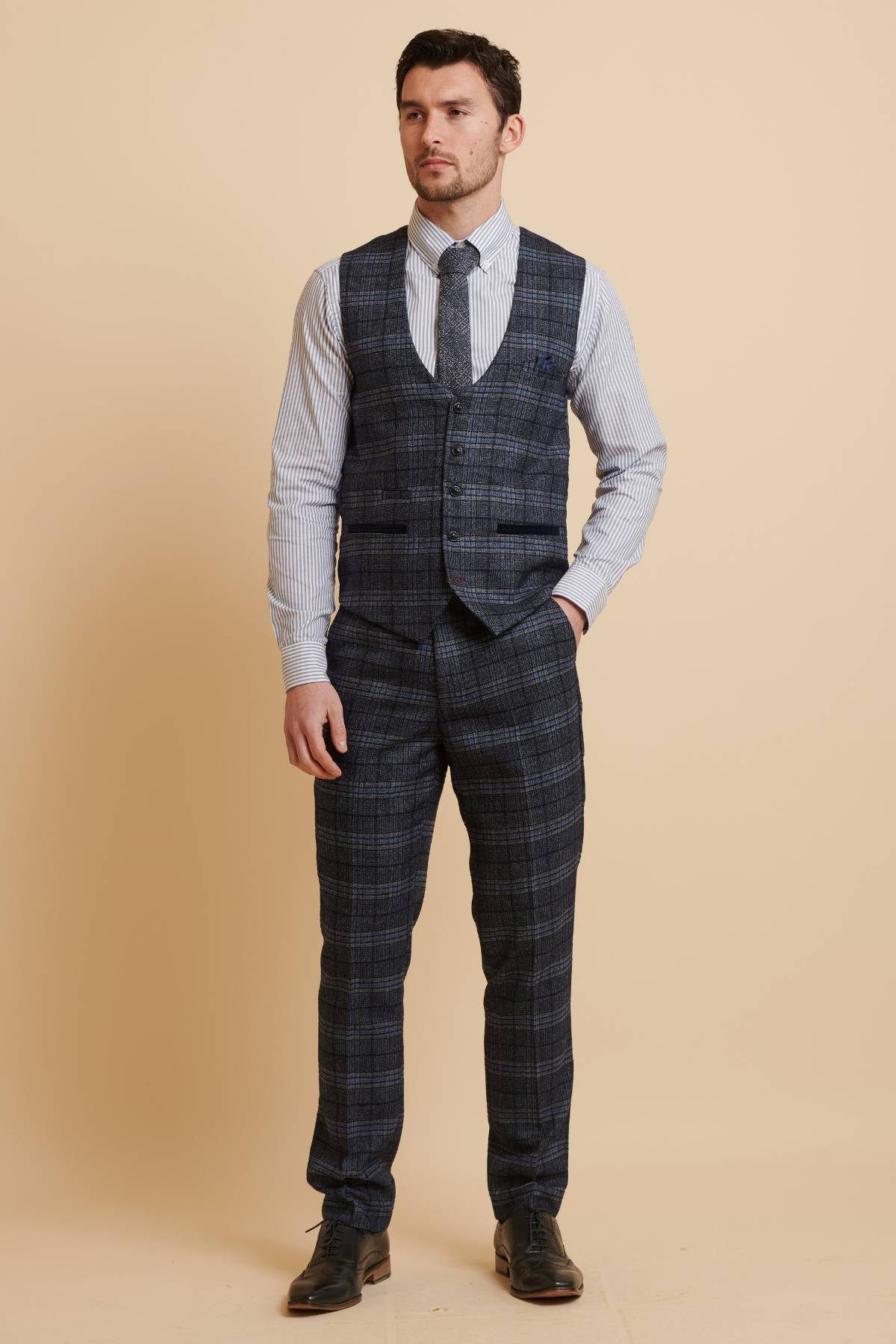 A man stands against a beige background wearing the Marc Darcy ANDY blue single breasted check waistcoat, styled with a blue plaid suit, white and blue striped shirt, dark tie, and black shoes. He looks forward with arms relaxed at his sides.