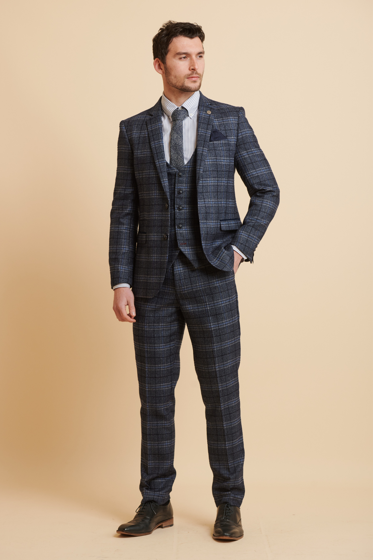 James Tarkowski models the ANDY Blue Check Suit from The Everton Collection by Marc Darcy against a beige background, paired with a white shirt, striped tie, and black dress shoes. He gazes slightly right with a neutral expression.