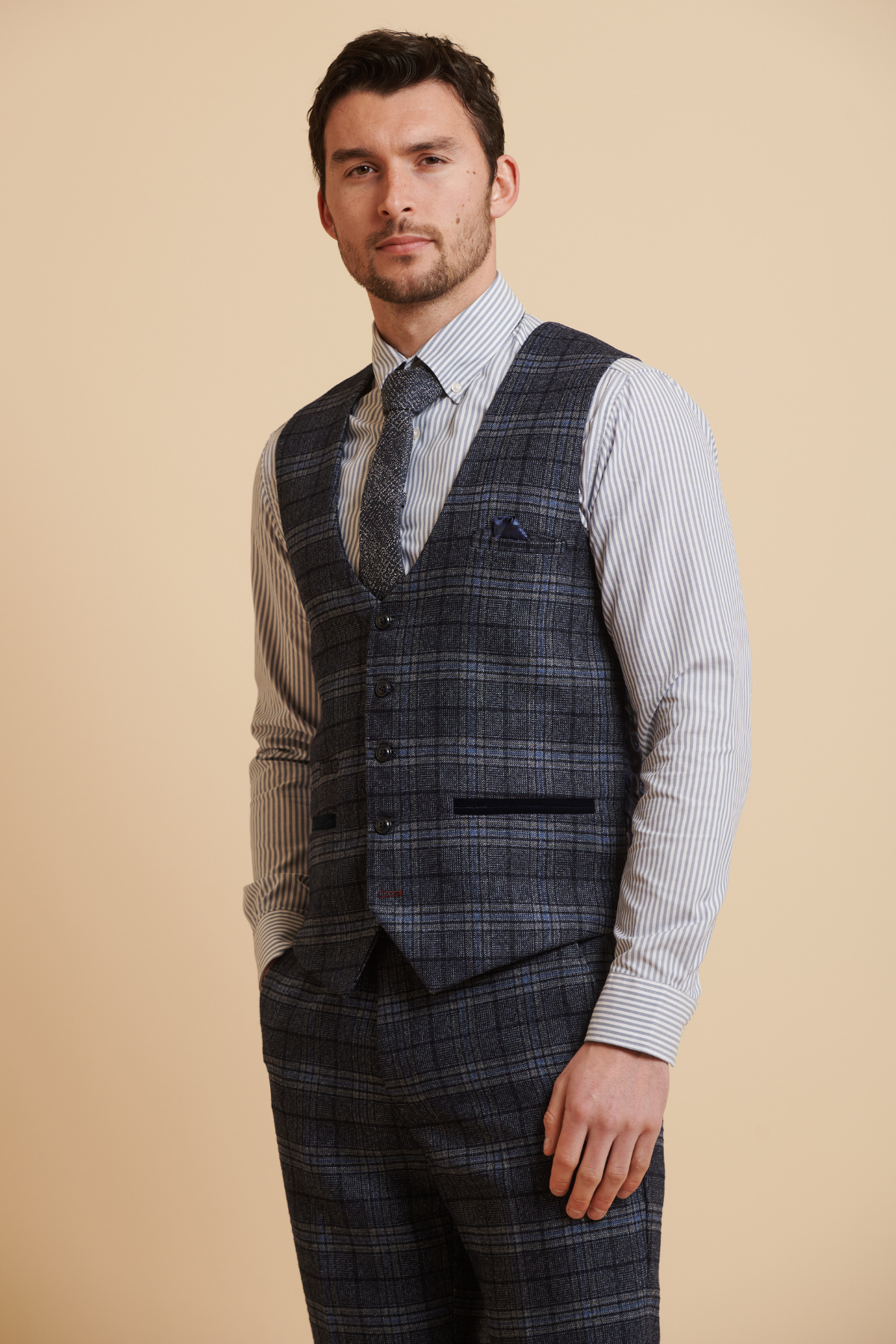 A man wearing the Marc Darcy ANDY Blue Single Breasted Check Waistcoat over a light striped shirt and matching tie stands confidently against a beige background.