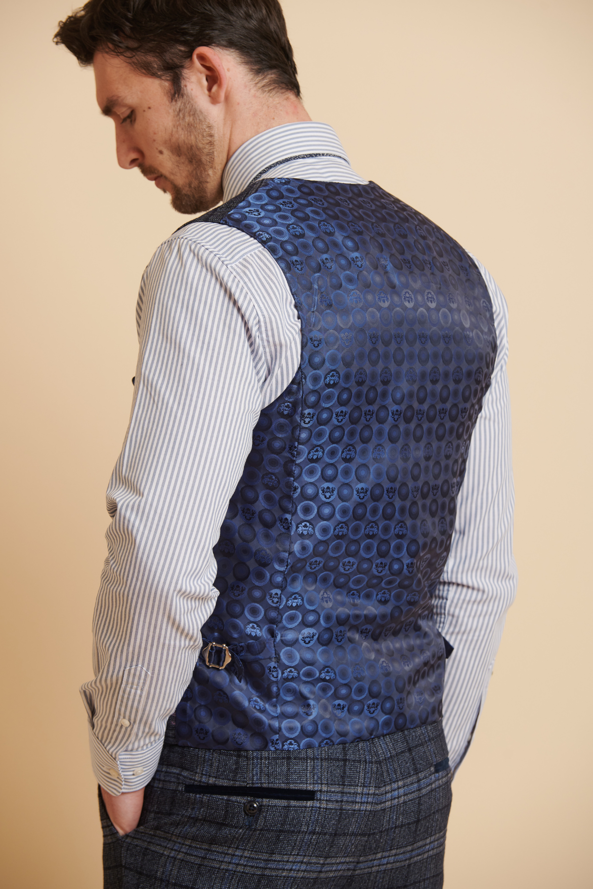 A man wears the ANDY - Blue Single Breasted Check Waistcoat by Marc Darcy over a striped shirt and plaid trousers, hands in pockets, creating a bold mix of formalwear and patterns.