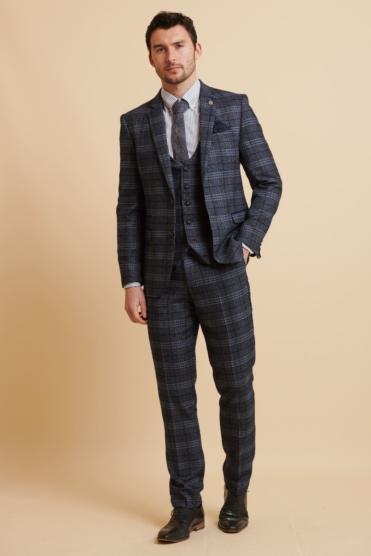 A man wears the Marc Darcy ANDY Blue Check Three Piece Suit with a white striped shirt, patterned tie, and pocket square, standing with his hand in his pants pocket.
