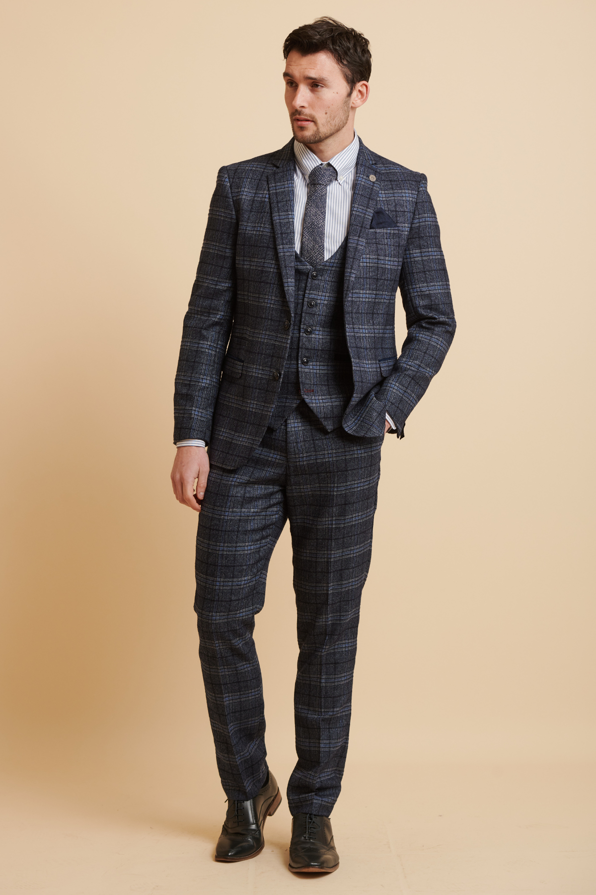 James Tarkowski models the ANDY Blue Check Suit from The Everton Collection by Marc Darcy, standing against a beige background in a slim fit three-piece suit with a striped shirt and tie, looking to the side with one hand in his pocket.