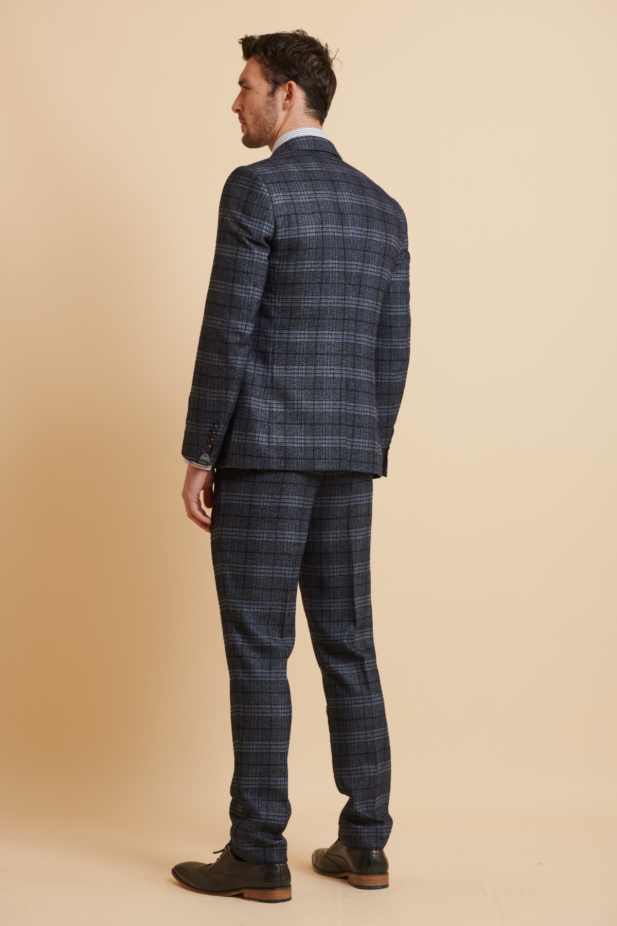 James Tarkowski wears the ANDY Blue Check Slim Fit Suit with matching pants by Marc Darcy from The Everton Collection, paired with black shoes, standing against a beige background.