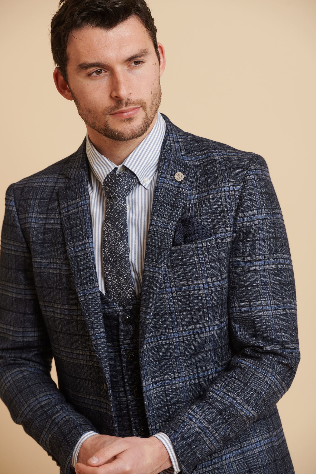 James Tarkowski wears the ANDY Blue Check Suit from The Everton Collection by Marc Darcy, paired with a striped shirt, patterned tie, and pocket square. He stands against a beige background, looking to the side with hands loosely clasped.