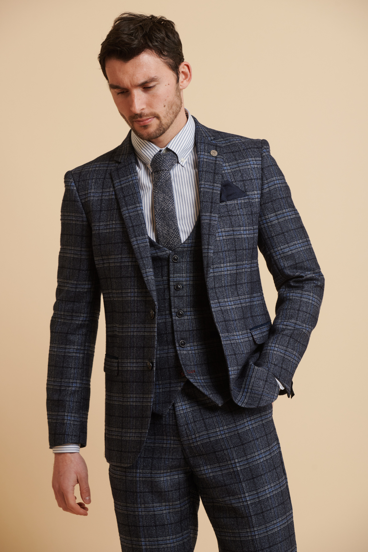 James Tarkowski wears the ANDY Blue Check Suit from The Everton Collection by Marc Darcy, styled with a striped shirt, grey knit tie, and navy pocket square—ideal for modern men's formalwear against a beige backdrop.