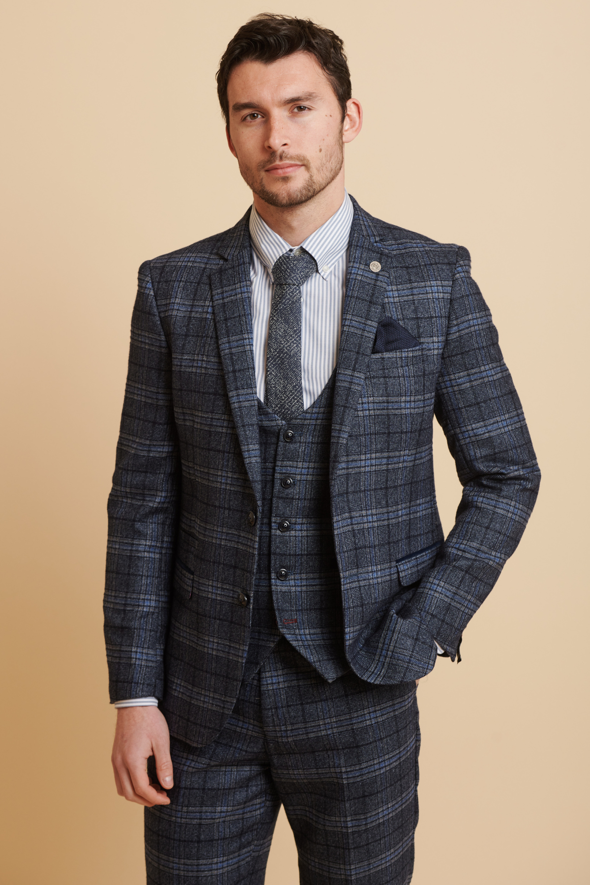 James Tarkowski wears the ANDY Blue Check Suit from The Everton Collection by Marc Darcy—styled with a matching vest, striped shirt, and tie—posing against a beige background with one hand in his pocket.