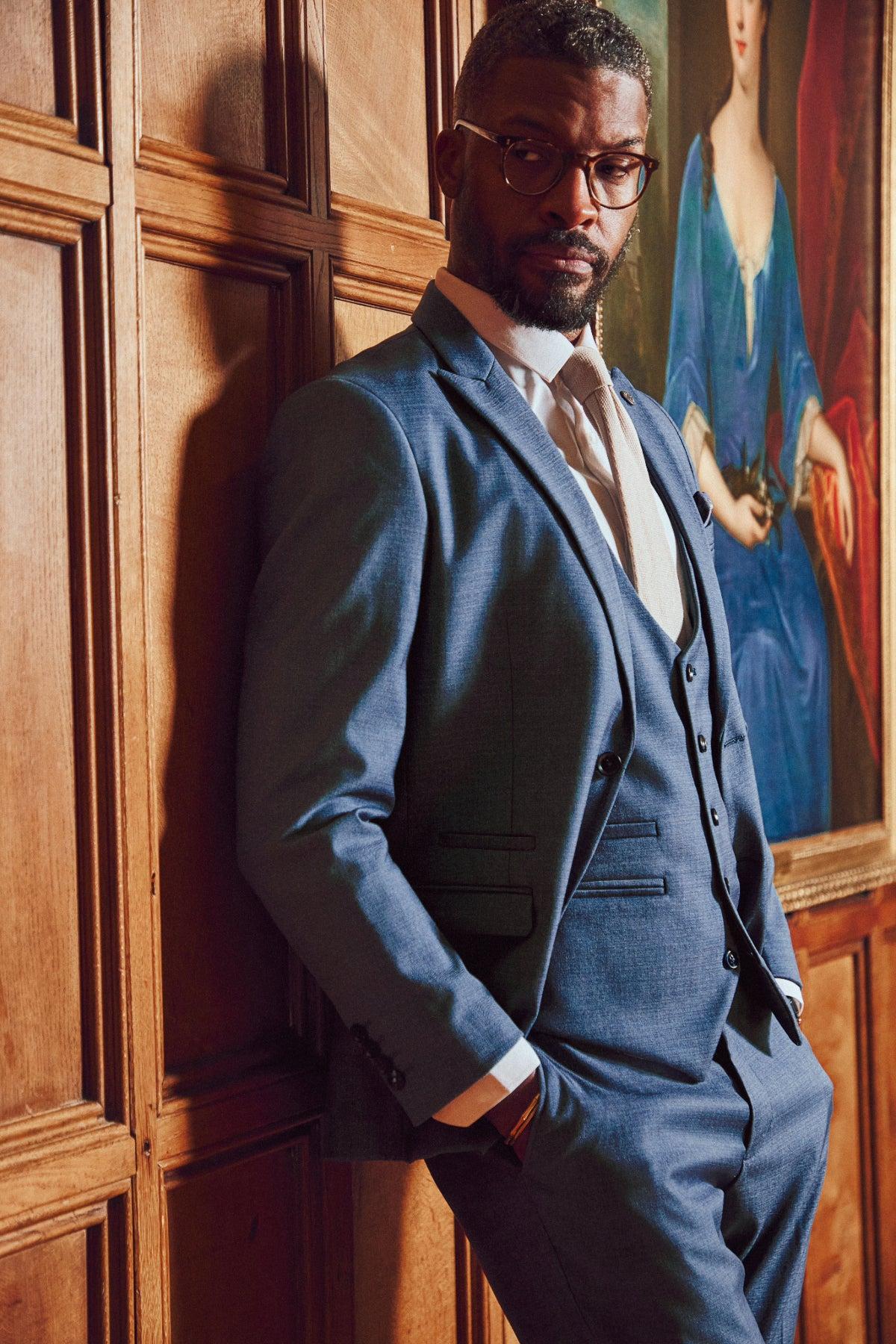 A man in the Marc Darcy FINLEY teal blue three piece suit with glasses leans against a wooden paneled wall beside a classic portrait of a woman in historical dress.