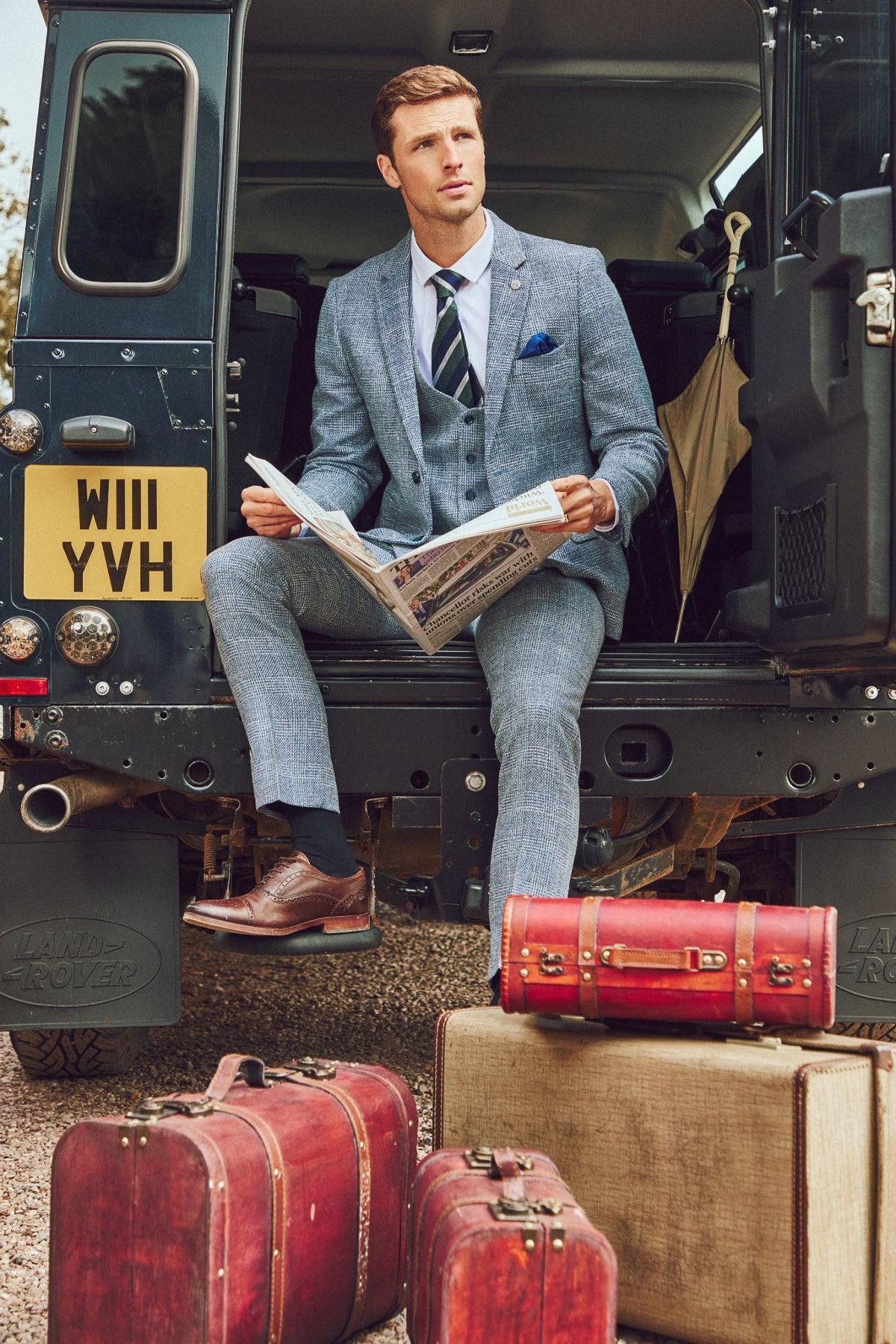 A man in a marcdarcy KRIS Sky Blue Tweed Three Piece Suit sits in the back of a Land Rover reading a newspaper, surrounded by vintage suitcases.