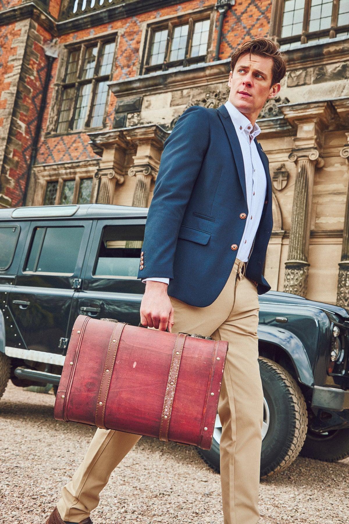 A man in a marcdarcy MAX Navy Blue Blazer with Contrast Buttons, white shirt, and tan pants walks outside a brick building carrying a vintage red leather suitcase and wearing a camouflage pocket square, with a dark SUV behind him.