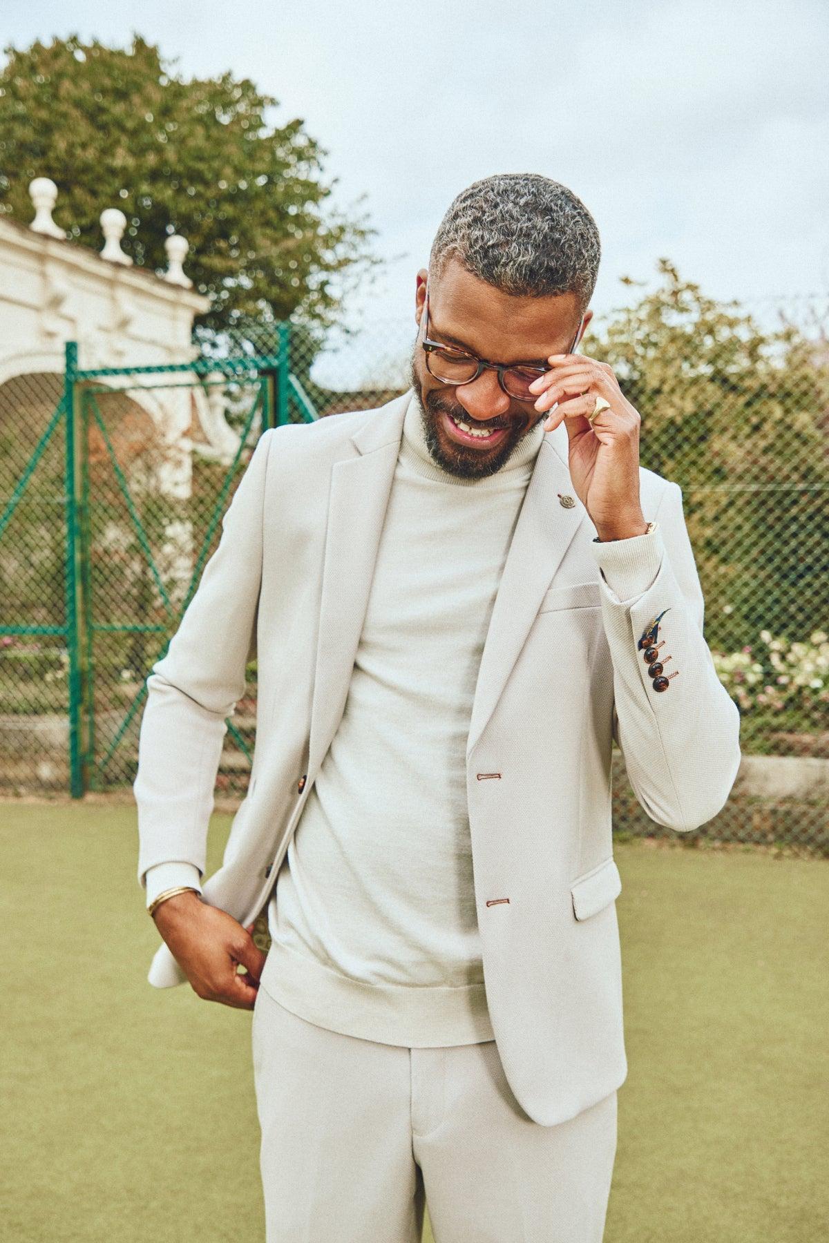 A man with short, curly gray hair and a beard smiles while wearing glasses and the Marc Darcy HM5 - Stone Tailored Two Piece Suit. He stands outdoors on a green surface near a fence and garden, adjusting his glasses.