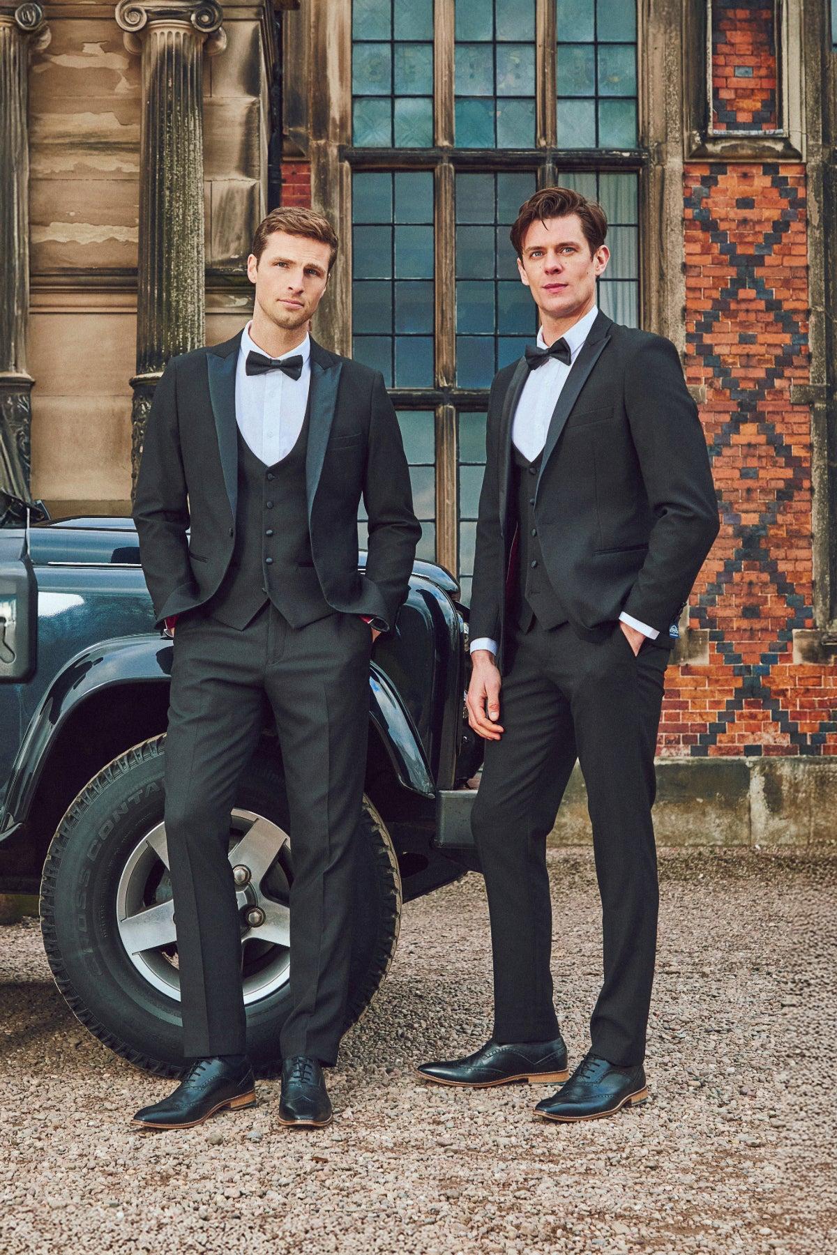 Two men wear marcdarcy DALTON Black Tux Lapel Diamond Three Piece Suits and bow ties, posing by a black car and a historic building—one leaning casually on the vehicle, the other standing with hands at his sides.
